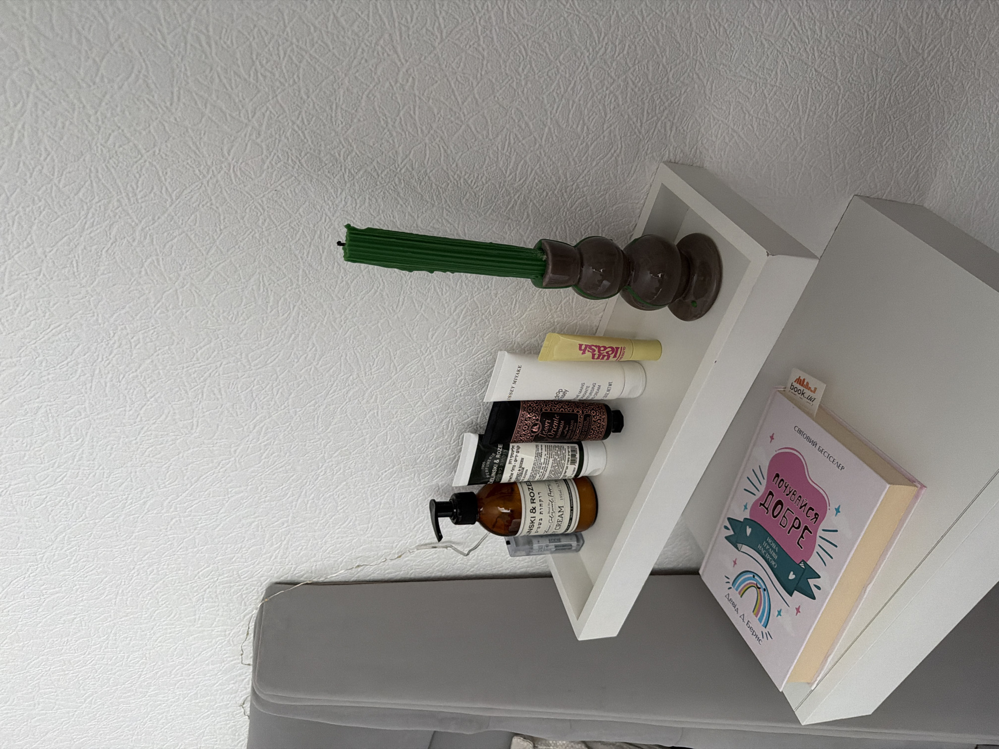 The image depicts a close-up, eye-level shot of two white floating shelves mounted on a white textured wall. On the upper shelf, from left to right, are a small clear object, a brown pump bottle with text on it, a white tube with black text, another white tube with black text, a small yellow tube, and a dark brown candle holder with a green candle. On the lower shelf, a pink book with colorful illustrations and text is placed horizontally, with its spine facing towards the viewer. The background is a plain white textured wall. There are no people visible in the image. The lighting suggests an indoor setting, possibly a bedroom or bathroom, with soft, diffused light. The scene is tidy and suggests a personal space. There is no information available in the image to determine the location in the city of Kyiv, Ukraine.