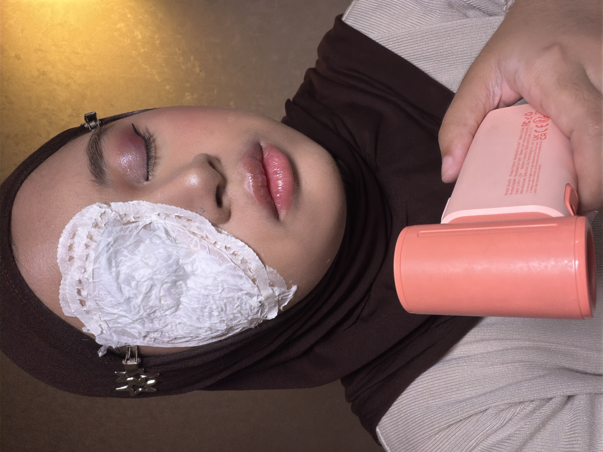 A close-up, angled shot shows a person in a brown hijab lying down, their face the central focus. A white, crumpled tissue or cloth is positioned over their left eye, held in place by clips. The person's right eye is closed and adorned with glittery, pink eyeshadow and black eyeliner. Their lips are glossy and pink. To the right of the person's face, a hand holds a pink, cylindrical device.
The background is partially gold, and the person is wearing a light gray shirt under the hijab. A sticker or label is visible on the back of the pink device, showing various symbols and text.