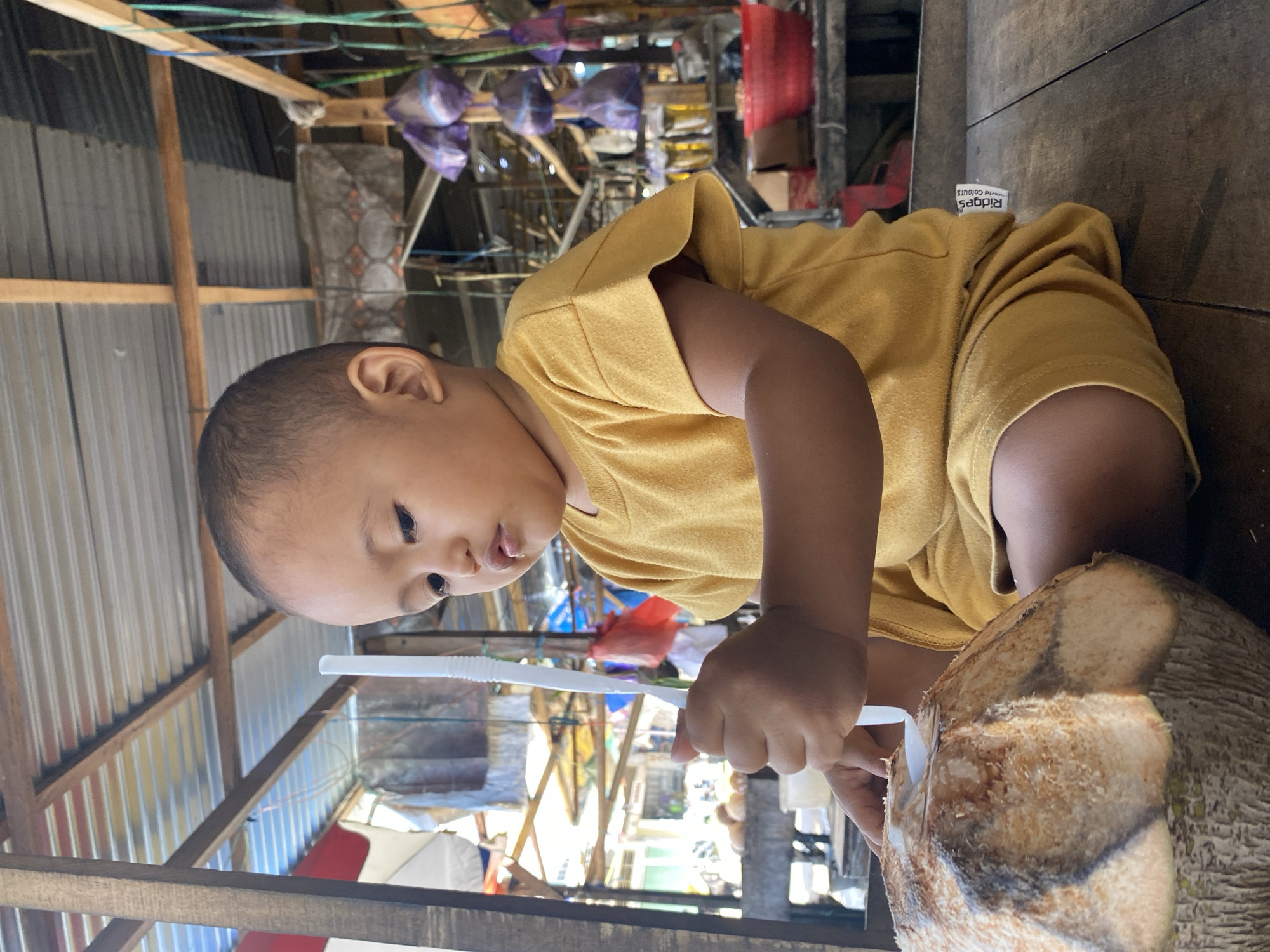A young child, likely a toddler, wearing a yellow t-shirt and shorts, is seated on a wooden surface at what appears to be an outdoor market stall in Ta', Indonesia. The child is actively engaged with a large, freshly opened coconut, using a white plastic straw and spoon to access its contents. The background reveals the typical market environment with corrugated metal roofing supported by wooden beams, shelves displaying various items in plastic bags, and a glimpse of the surrounding street with other stalls and structures. The child's focused expression and the casual setting suggest a moment of everyday life.