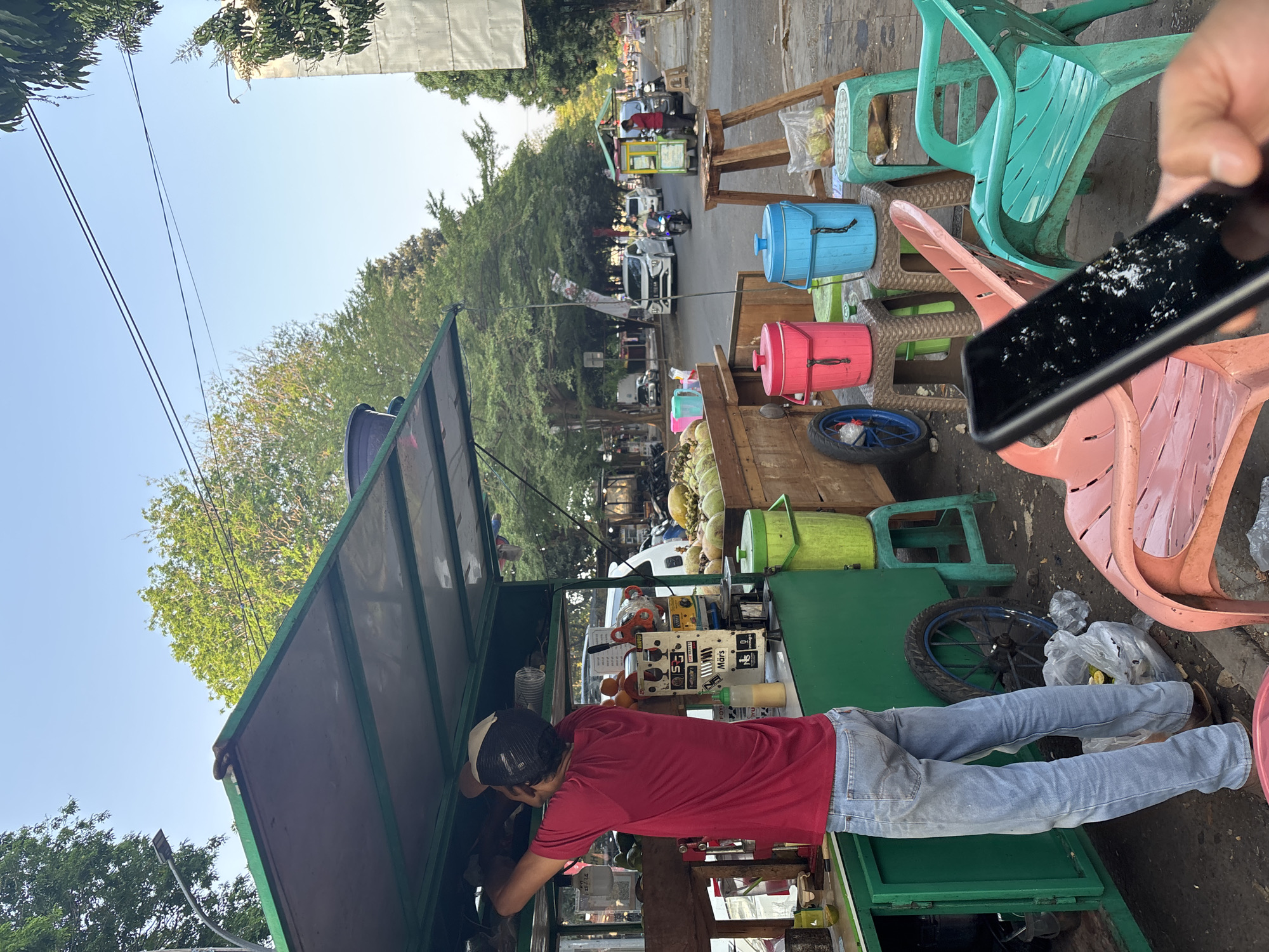 The image captures a street-side juice stand in Cirebon, Indonesia. A man in a red t-shirt and light blue jeans is working at the stand, which is adorned with a green canopy. Various items are visible, including large green coconuts stacked in a wooden crate, a juice dispenser, plastic buckets in pink and blue, and several colorful plastic chairs. The stand is located on the side of a street with trees lining the sidewalk and cars and motorcycles driving by in the background. The scene is illuminated by natural daylight, suggesting it is either morning or late afternoon.