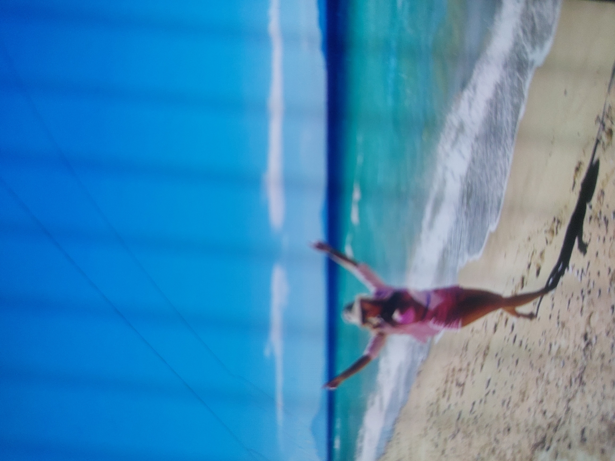 A woman in a pink swimsuit is on a beach in Tsarevo, Bulgaria. She stands on the sand with her arms outstretched. The sea is turquoise and laps the shore with white waves. The sky is blue. The image is oriented sideways.