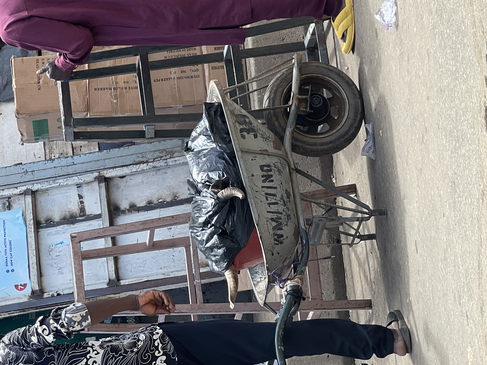 The image shows a ram carcass being transported in a wheelbarrow in Abuja, Nigeria. The ram carcass is wrapped in a black plastic bag. The wheelbarrow is old and has the letters "UNI" partially visible on its side. Two people are visible in the image. One is wearing black pants and a black and white patterned shirt, and the other is wearing purple pants. They are standing near a pile of boxes on a metal frame. The boxes appear to contain whiteboard markers and are labeled "MADE IN NIGERIA." The setting appears to be outdoors, possibly on a street or near a market.