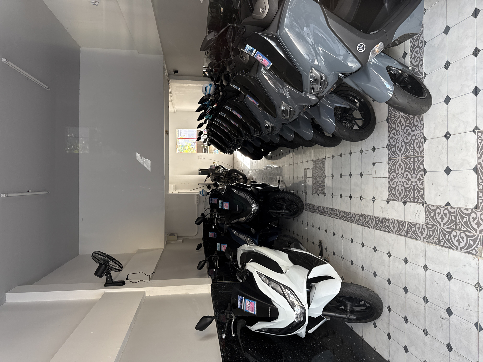 This is a medium shot of a motorcycle showroom in Phuket City Municipality, Thailand. The showroom is indoors, with white walls and a grey ceiling. The floor is tiled with a white and grey patterned design. There are several motorcycles parked in rows, facing the viewer. The motorcycles are mostly dark-colored scooters, with one white scooter in the foreground on the left. Some of the motorcycles have price tags displayed on them. A black oscillating fan is mounted on the left wall, plugged into an electrical outlet. A fluorescent light fixture is visible on the ceiling. Sunlight streams in from an open doorway at the far end of the showroom. The scene is clean and orderly, suggesting a commercial establishment. There are no people visible in the image.