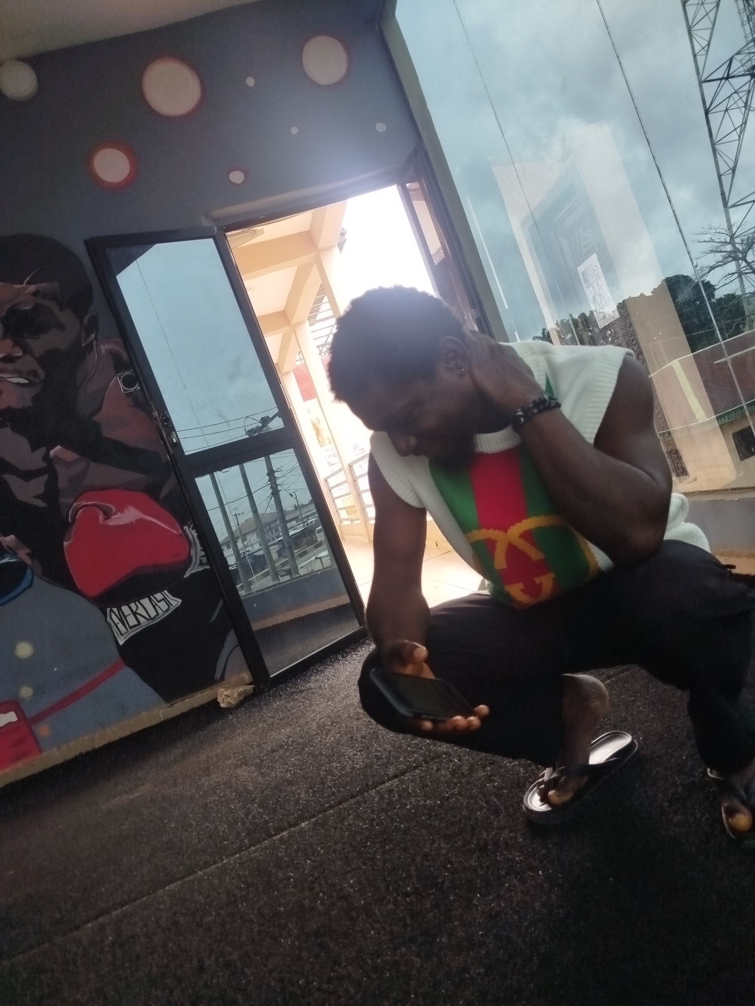 A man is captured in a crouching position, looking down at his phone, in what appears to be an indoor gym in Uselu, Nigeria. He is wearing a white sleeveless sweater with a green, red, and gold striped pattern, and black pants. His left hand is holding a black smartphone, while his right hand is touching the back of his neck. He is wearing black flip-flops. Behind him, a large mural of a boxer is visible on the wall, with the word "EVERLAST" written on the boxer's shorts. The gym has a large glass door or window, through which the sky and some buildings are visible. The floor is covered with dark, textured material, likely rubber flooring common in gyms. The lighting suggests it is daytime, with light streaming in from the glass area.
