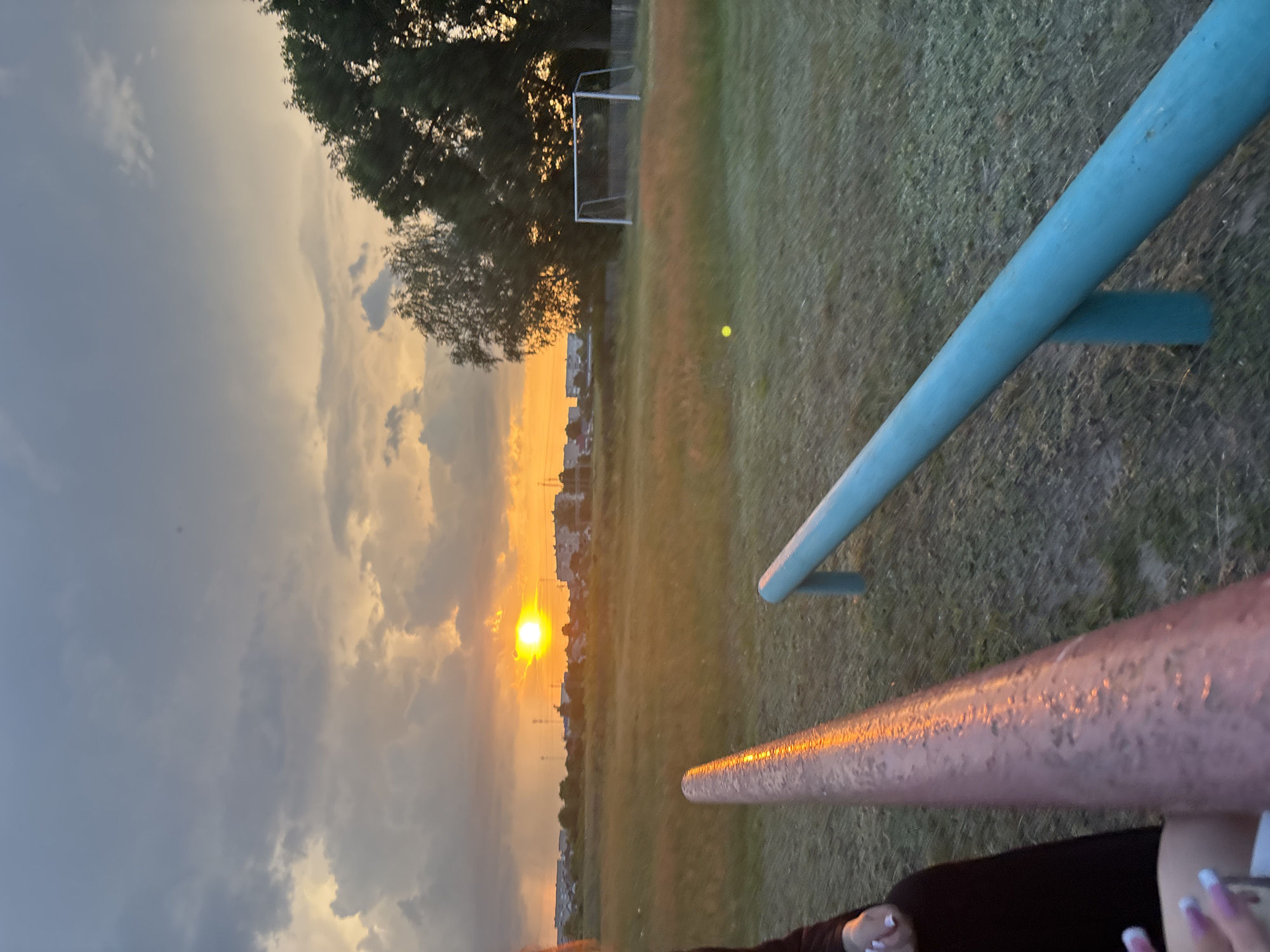 The image shows a sunset over a grassy field in the city of Сароцкі сельскі Савет, Belarus. The sun is setting behind a cluster of buildings, casting a warm orange glow across the sky and the landscape. In the foreground, two metal bars, one pink and one blue, are visible. A person's hand, with manicured nails, is visible near the pink bar. In the background, there is a soccer goal and a few trees. The overall scene depicts a peaceful, outdoor setting during twilight.