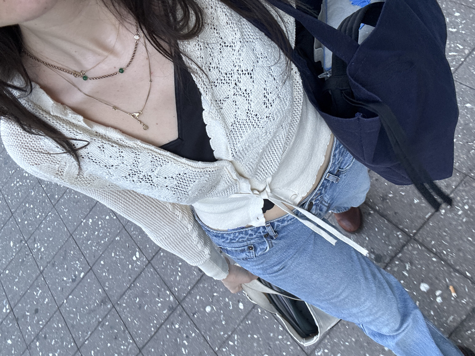 An overhead shot captures a person from the chest down, showcasing their casual daytime outfit and accessories while standing or walking on a paved surface in what appears to be an urban environment.
The subject is wearing a light-colored, possibly cream or off-white, open-knit cardigan with a distinct lacy pattern, tied loosely at the front. Beneath the cardigan, a black bralette or camisole is visible. They are paired with light wash, high-waisted blue denim jeans. On their feet, one brown shoe, possibly a loafer or boot, is partially visible on the right side of the frame.
Several accessories adorn the outfit. Layered gold-toned necklaces are prominent: one shorter chain features small green beads, while a longer chain ends with a distinct clasp or charm. The person carries two bags: a dark navy blue tote bag is slung over their right shoulder, and a light-colored (possibly beige or cream) tote bag is held in their left hand. Long, dark hair frames the left side of the image.
The setting is an outdoor area with grey square pavement tiles, speckled with white and light grey, suggesting concrete or asphalt with aggregate. The lighting is bright and even, indicating daytime. No text is visible in the image. The overall impression is a relaxed, stylish urban look.