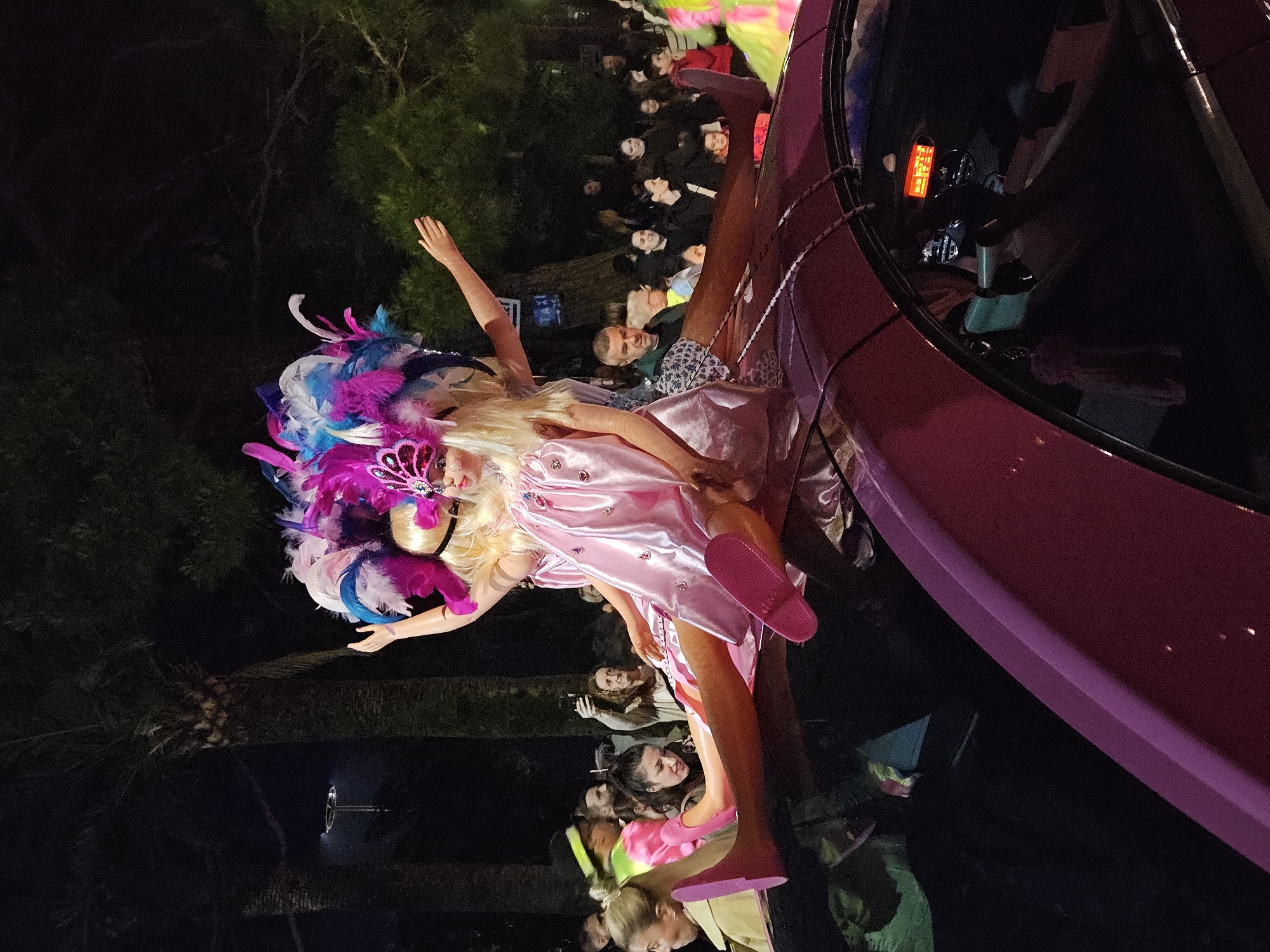 The image shows a parade at night. A large pink vehicle with a mannequin dressed as a doll mounted on top moves through the crowd. The mannequin has blonde hair and is wearing a pink dress and a feathered headdress in shades of pink, white, and blue. People are standing along the sides of the road watching the parade. The location is Unknown, Unknown.