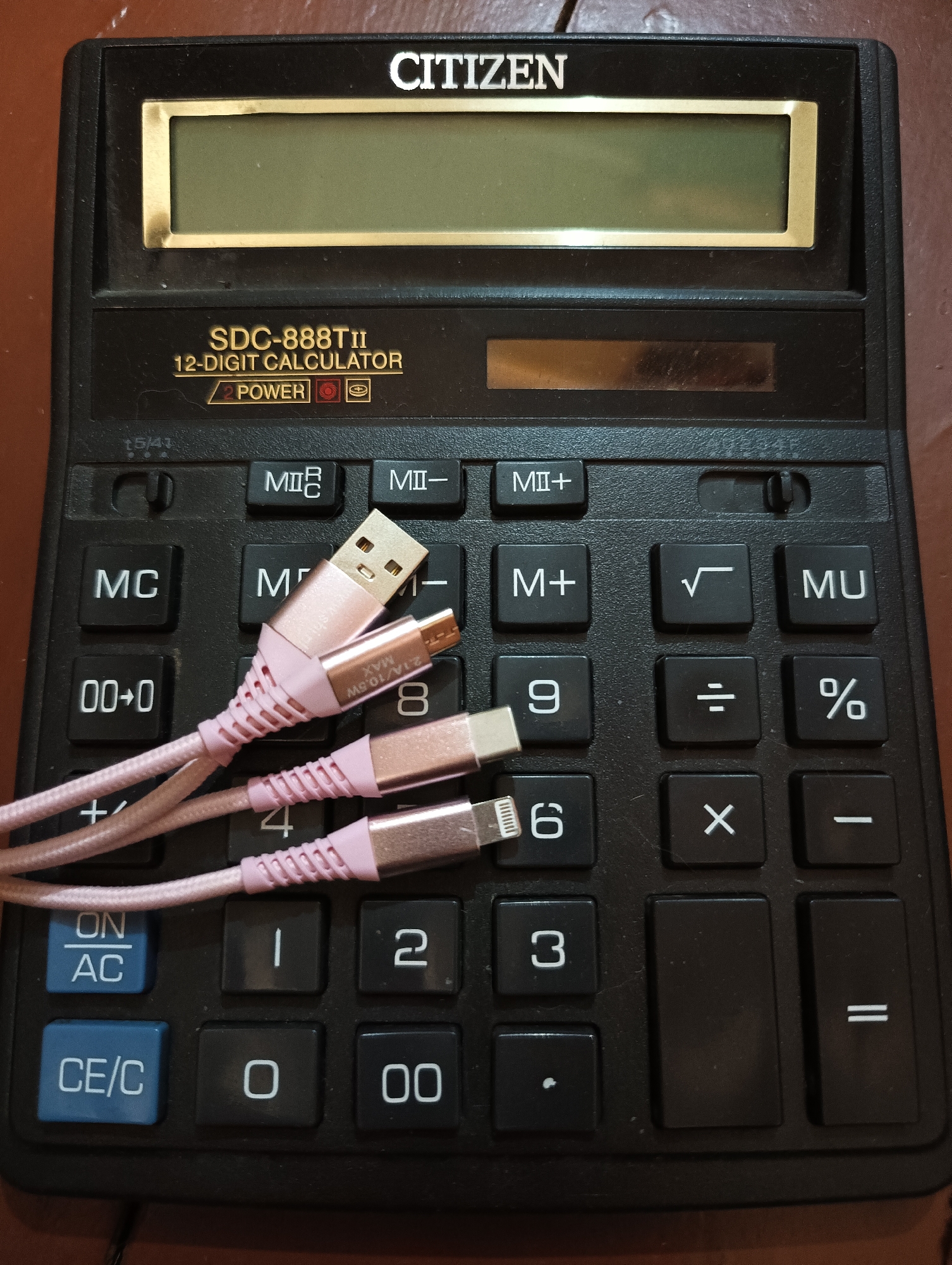 An overhead photograph displays a black Citizen SDC-888TII 12-digit calculator situated on a brown, possibly wooden, surface in Biliaivka, Ukraine. The calculator has a gold-trimmed LCD screen and a small solar power strip. Key labels visible include "CITIZEN", "SDC-888TII", "12-DIGIT CALCULATOR", and "2 POWER". A partially visible "15/41" is also present.
Laid across the calculator's numeric and function keys are three pink USB cables. These include a USB-A to USB-C cable, a USB-A to Lightning cable, and a USB-A to Micro-USB cable. On one of the cable connectors, the brand "wushin" and specifications "2.1A/10.5W MAX" are discernible.
The calculator's keypad features standard arithmetic operations and memory functions, such as "ON/AC", "CE/C", "MC", "M", "M+", "MI+", "MI-", "MU", "√", "%", "+", "-", "X", "÷", "=", along with numbers "0" through "9", "00", ".", and "00+0". There is no active event or human subject. The scene is a static arrangement of electronic accessories on a household surface, without specific time of day or weather indications, and no discernible emotions are conveyed.