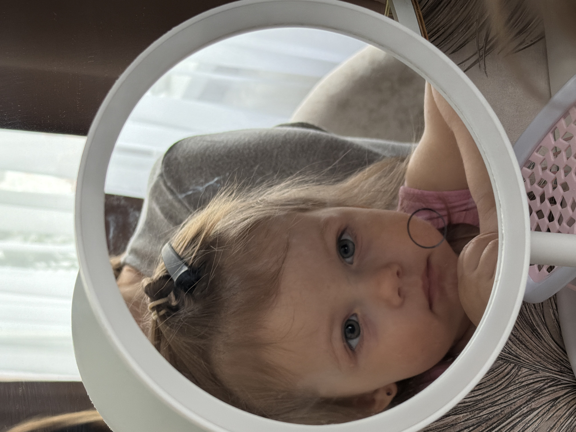 A close-up image shows the reflection of a young child in a round, white-framed mirror or ring light. The child, with light brown hair and prominent blue-gray eyes, gazes directly into the mirror with a neutral and curious expression. A small black hair clip is visible in her hair on the left side of her head. She is wearing a pink top, and a faint, thin black ring mark is visible on her face near her mouth.
The background reflected in the mirror suggests an indoor, domestic setting during daytime, with soft light coming from what appears to be a window or light source behind the child. A large, gray or tan cushion or soft furniture item is visible behind the child's head. To the right of the child's reflection, a pink, lattice-patterned plastic object is partially visible, possibly part of a toy or piece of furniture. The general composition is focused on the child's face within the circular reflection. No text is visible in the image.
