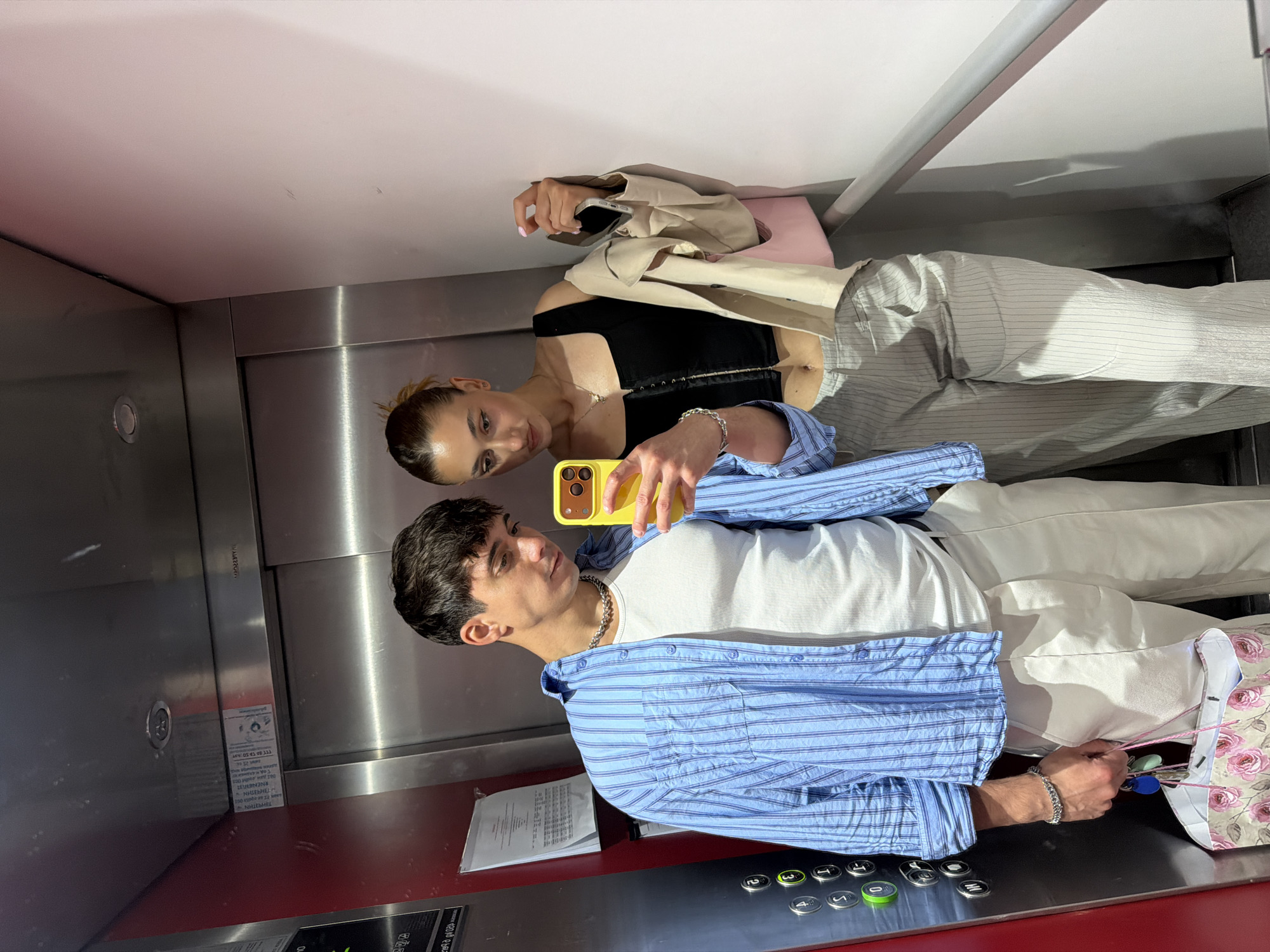 Two young individuals, a man and a woman, are captured in a mirror selfie inside an elevator in Sofia, Bulgaria. The image is rotated 90 degrees clockwise.
In the foreground, a young man holds a bright yellow smartphone to take the photo. He wears a white ribbed t-shirt layered under an unbuttoned blue and white striped long-sleeve shirt, paired with white pants. He accessorizes with a silver chain necklace and a silver chain bracelet on his left wrist. He holds a set of keys attached to a ring with various colored fobs in his right hand, alongside a floral-patterned bag.
Behind him and slightly to his left is a young woman. She wears a black top, light-colored vertically striped pants, and has a beige jacket draped over her left arm. A light-colored handbag rests next to her. Her hair is pulled back, and she wears a simple gold necklace. Both individuals are looking towards the camera/phone reflection.
The elevator interior features brushed metal walls and a red floor. A control panel with several numbered buttons (including 2, 3, 4, 5, and T) and a prominent green button is visible on the right. Papers are placed on a ledge below the control panel. On the left metal wall, a sticker with Cyrillic text and numbers like "49 13 50" is partially visible, likely an elevator inspection or information notice. The ceiling appears white. The overall impression is an everyday moment captured candidly.