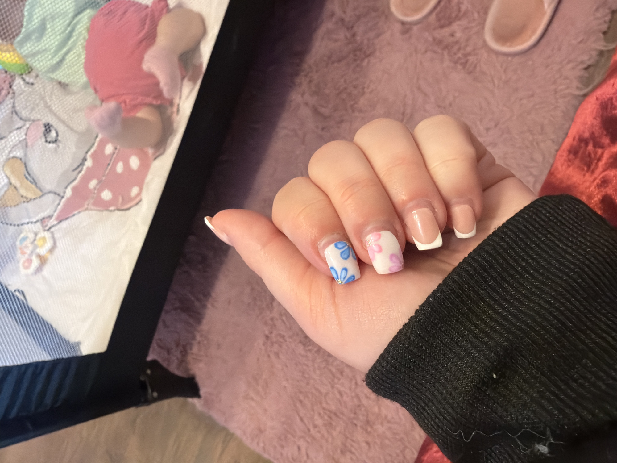 A hand with manicured nails rests on a light pink, soft, plush surface. The nails on the index and middle fingers have floral designs (blue and pink, respectively), while the other nails have a French manicure style. The person is wearing a black sweater with a ribbed texture. In the background, there are a pair of light pink slippers and a baby doll. Part of a baby cot is also visible on the left side. There is no indication that this image was taken in Varna, Bulgaria.