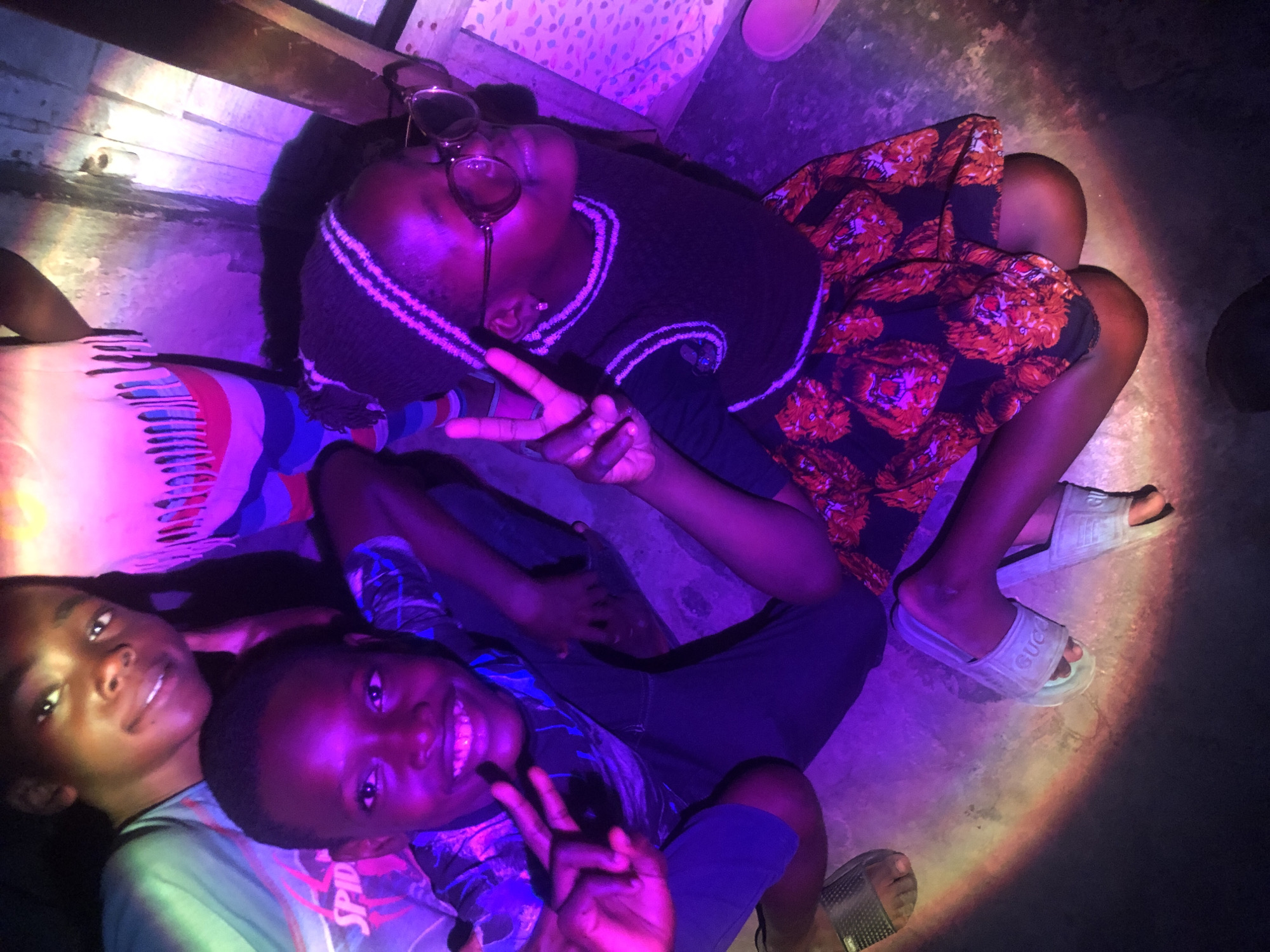 In the city of Ado, Nigeria, under purple-hued lighting, two young boys and a young girl are captured in an upward-facing shot. The boy in the foreground, on the left, with dark skin and short black hair, is smiling broadly and making a peace sign with his left hand. He wears a dark blue t-shirt with a graphic design and blue shorts. To his right, a young girl with dark skin, wearing a knitted beanie and glasses, also makes a peace sign with her left hand. She has on a dark top with stripes on the collar and a black skirt adorned with red floral patterns. Behind them, another boy with dark skin, smiling with visible teeth, is partially visible. The environment appears to be indoors, with a concrete floor and a wall with wooden planks and some colorful, possibly painted or tiled, sections illuminated by the purple light. The lighting creates a shadow on the floor in the foreground, resembling a rainbow effect.