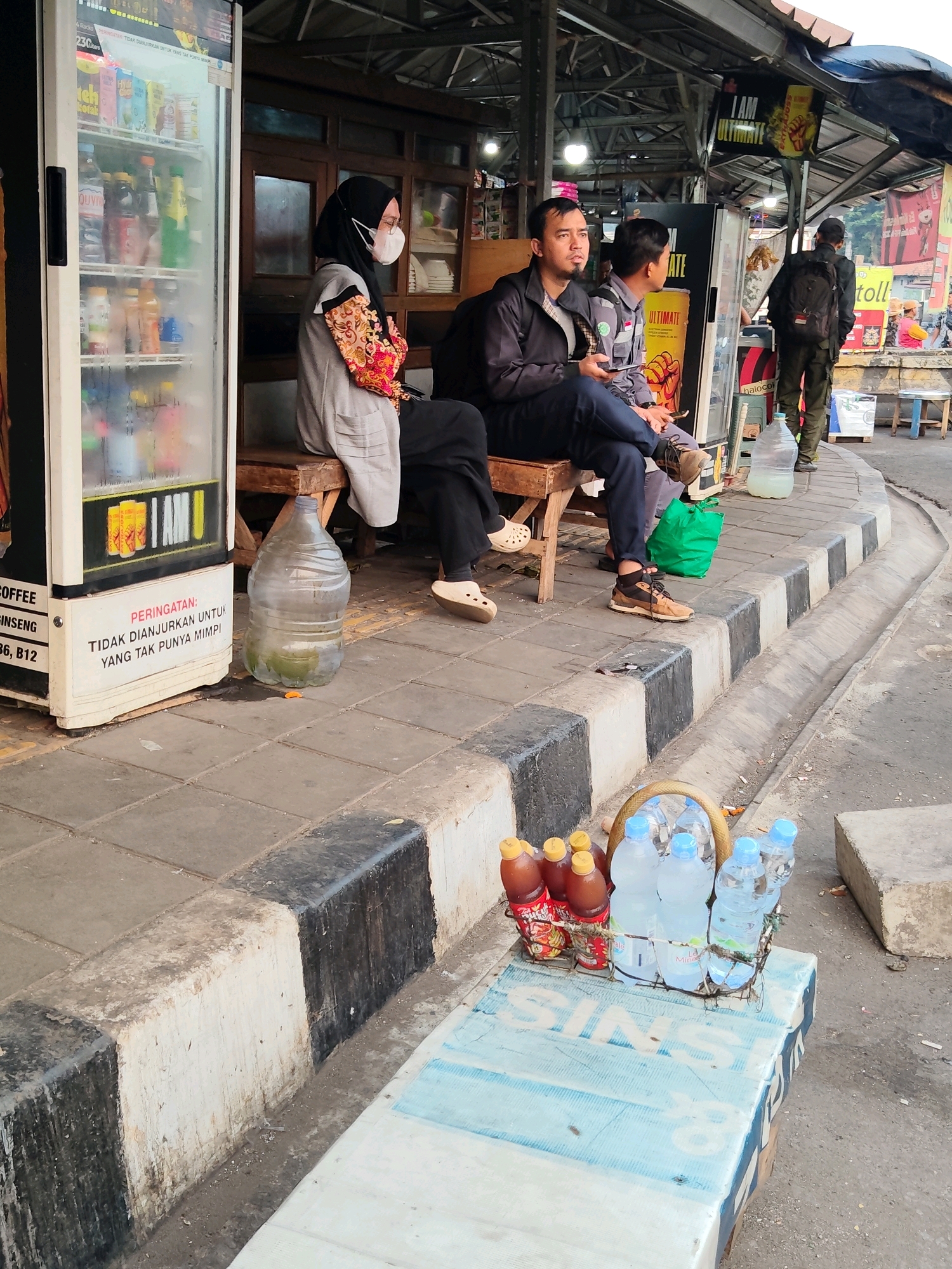 In Cikampek, Indonesia, a street scene unfolds near a roadside food stall. The foreground features a wooden surface displaying a basket containing bottled water and bottled beverages, likely for sale. To the left, a prominent white refrigerator unit, possibly a drink cooler, stands with its door open, revealing rows of bottled drinks. Next to it, a large transparent water jug sits on the sidewalk.
Further into the scene, several individuals are seated on a wooden bench situated outside the covered area of the food stall. One person, wearing a black hijab and a mask, is seated on the far left, their arms crossed. Next to them, two men are seated, one in a dark jacket and the other in a grey uniform, both looking forward. Behind them, a man wearing a backpack is standing and looking away from the camera. The environment is a paved sidewalk with a curb, separating it from a road or pathway. A green bag is visible on the ground near the seated individuals. The overall impression is a casual gathering or waiting area associated with a local vendor.