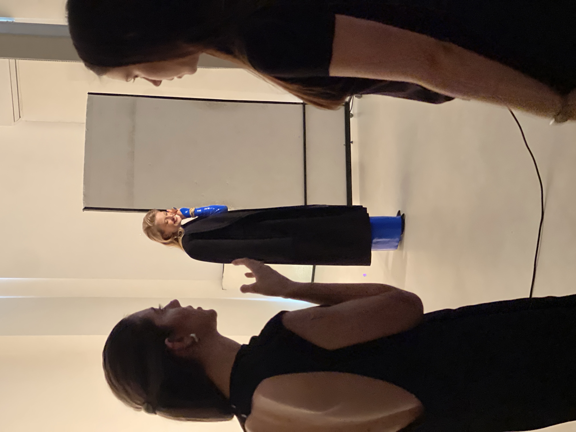In a dimly lit studio space with plain white walls, three individuals are present. In the foreground to the left, a woman with dark hair tied back and wearing a black open-back dress is seen in profile, facing a woman on the right. Her left hand is raised, fingers spread, in a gestural manner. To the right, another woman with dark hair, also seen in profile, is dressed in a black t-shirt and facing left. In the background, a blonde woman stands in front of a large white screen on wheels. She is wearing a black coat over bright blue trousers or a skirt, and her right hand is near her face, with a blue sleeve visible. The floor is a light, neutral color. The overall scene appears to be a photo shoot or a creative meeting. There is no discernible information in the image to suggest a specific location within the city of Kyiv, Ukraine.