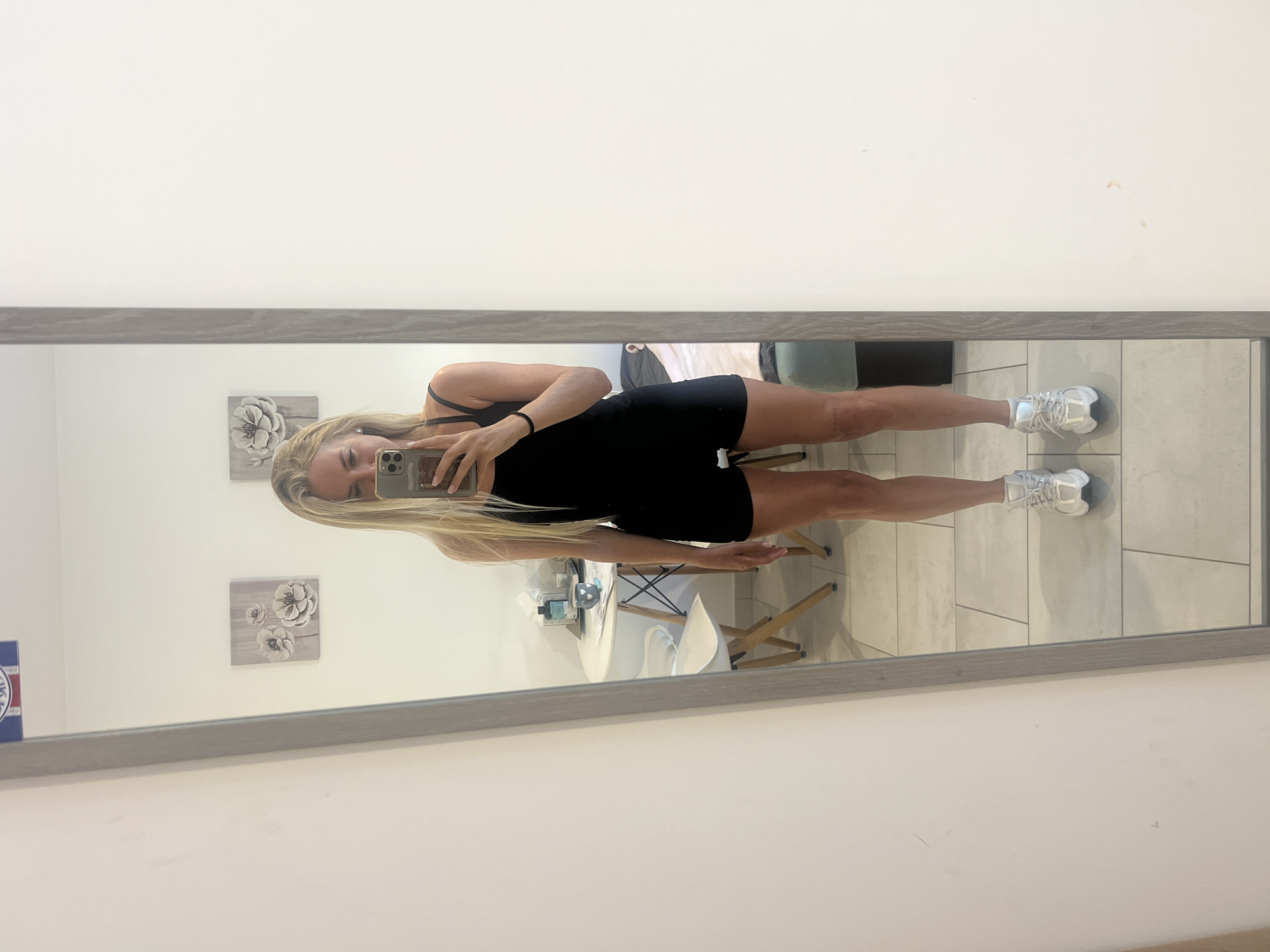 In Budva, Montenegro, a young woman with blonde hair stands in front of a full-length mirror, taking a selfie with her phone. She is wearing a black one-piece athletic outfit and white sneakers. The mirror reflects a room with tiled flooring, a small round table with chairs in the background, and two framed floral art pieces on the wall. The overall scene appears to be an indoor residential or hotel setting.