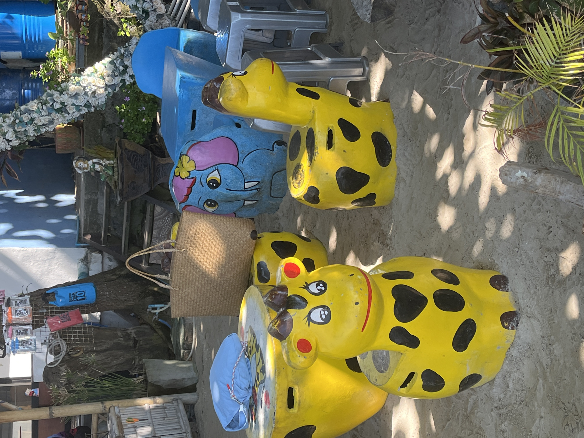 The image depicts a collection of objects in Ban Bang Khao, Thailand. There are blue and yellow painted animal-shaped seats, including elephants and giraffes. A woven bag sits near one of the elephant seats. Also visible are silver plastic chairs, blue barrels, various plants, a tree trunk with displayed merchandise (including a blue "10L DRY PACK"), and a bamboo structure. The ground appears to be sandy or gritty.