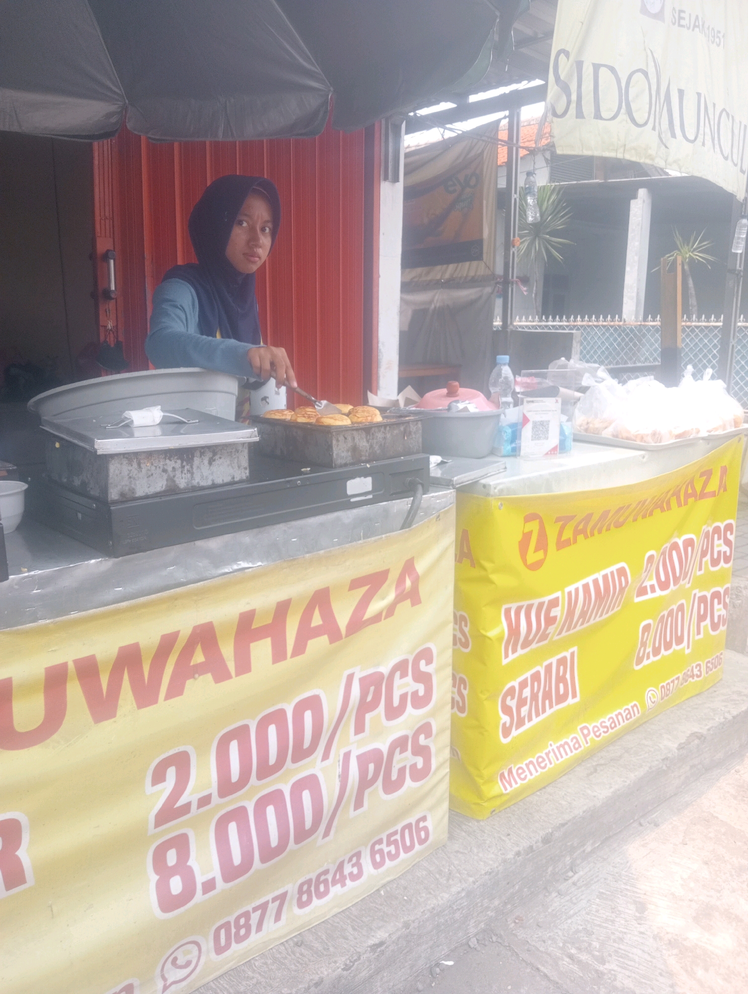 In a street-level setting in South Jakarta, Indonesia, a woman wearing a dark hijab and a blue long-sleeved shirt is preparing food at a food stall. She is facing the camera and holding a spatula over a tray of cooked, round food items, likely pancakes or crepes, situated on a flat-top grill. Behind her, a red corrugated metal wall forms the backdrop. The stall features a counter with various items including a large grey basin, a metal container, and plastic bags filled with food. Two yellow banners are prominently displayed. The banner on the left has red text reading "UWAHAZA" and "2.000/PCS" and "8.000/PCS". The banner on the right has red text reading "ZAMURAH KAZA", "KUE MAMIP 2.000/PCS", "SERABI", and "8.000/PCS". Below these prices, the banner also shows the text "Menerima Pesanan" (which means "Accepting Orders" in Indonesian) followed by a WhatsApp phone number and icons. A QR code and a bottle of water are visible on the counter. In the background, a chain-link fence and palm trees can be seen. The overall scene depicts a bustling street food vending operation.