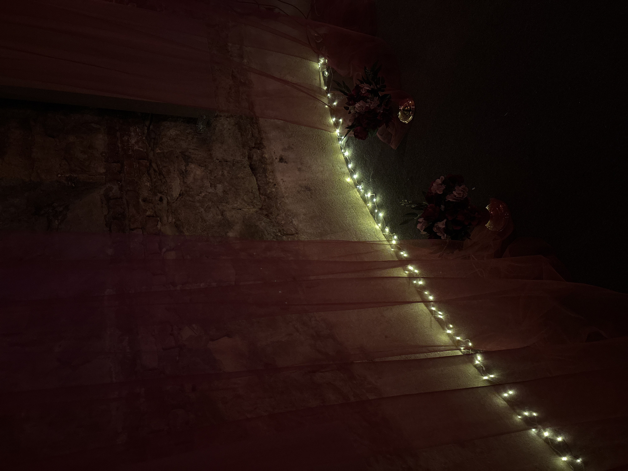 The image depicts a staircase draped with a sheer, reddish-pink fabric, illuminated by a string of warm white fairy lights. The staircase is constructed from stone or concrete. Two floral arrangements are present, one near the top of the stairs and another lower down. The background is dark. The location is likely Varna, Bulgaria.