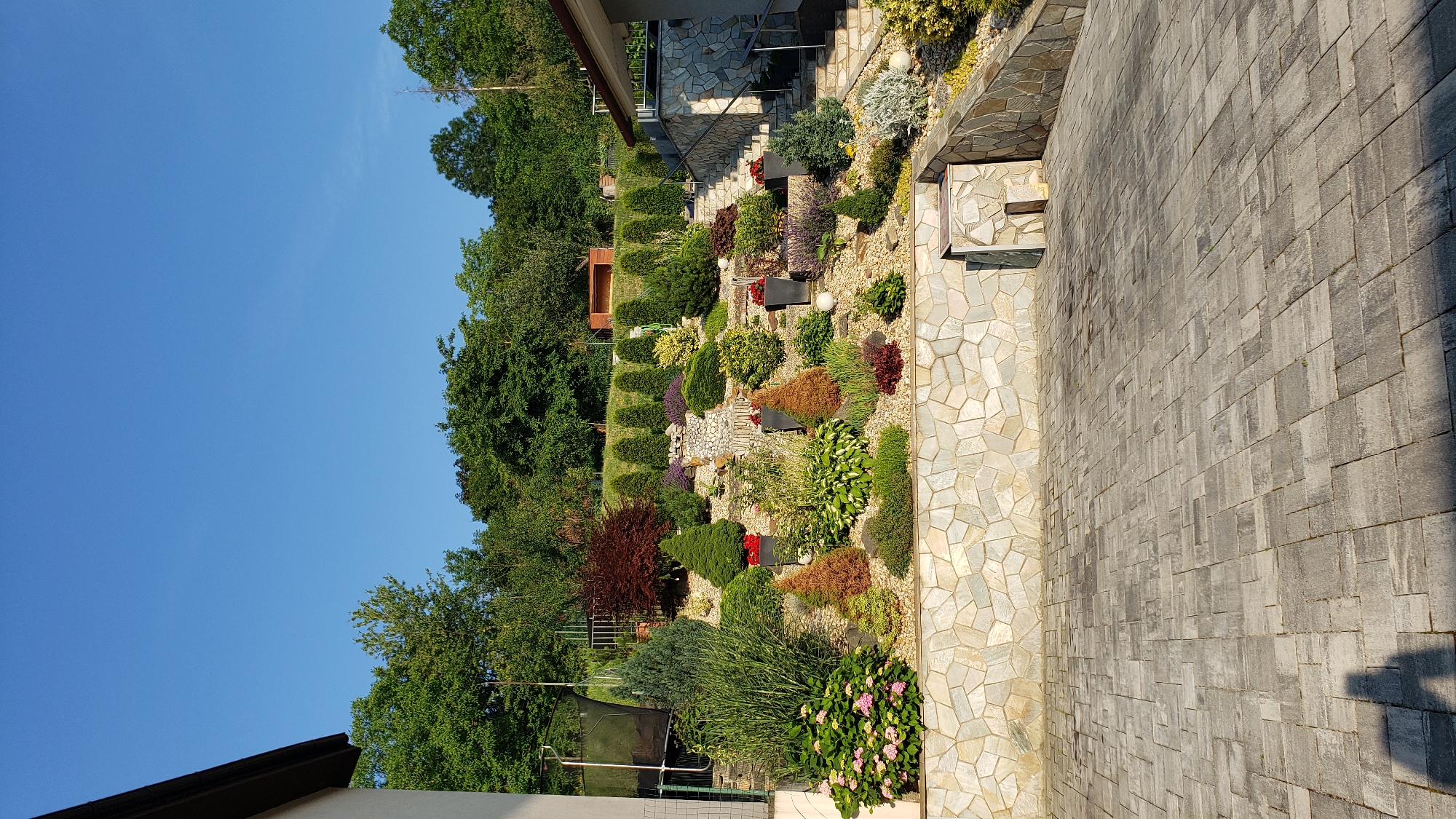 The image depicts a multi-tiered garden landscaping project on a hillside, likely part of a private residential property. The foreground is a paved patio area made of gray interlocking stones. A shadow of a person is cast on the patio. A stone retaining wall, constructed with irregularly shaped light-colored stones, forms the base of the garden. Above the wall, the garden features a variety of plants, including ornamental shrubs, evergreen trees, and flowering bushes, arranged in a terraced fashion. A small, decorative waterfall is integrated into the landscaping. A set of stone stairs leads up the right side of the hillside, alongside another section of the stone retaining wall, toward a building entrance. The background consists of a dense line of trees and a clear blue sky. The overall scene suggests a well-maintained outdoor space designed for aesthetic appeal and possibly recreation. Without further information, the exact location in Prešov, Slovakia cannot be determined, but the setting is characteristic of a suburban or rural residential area.
