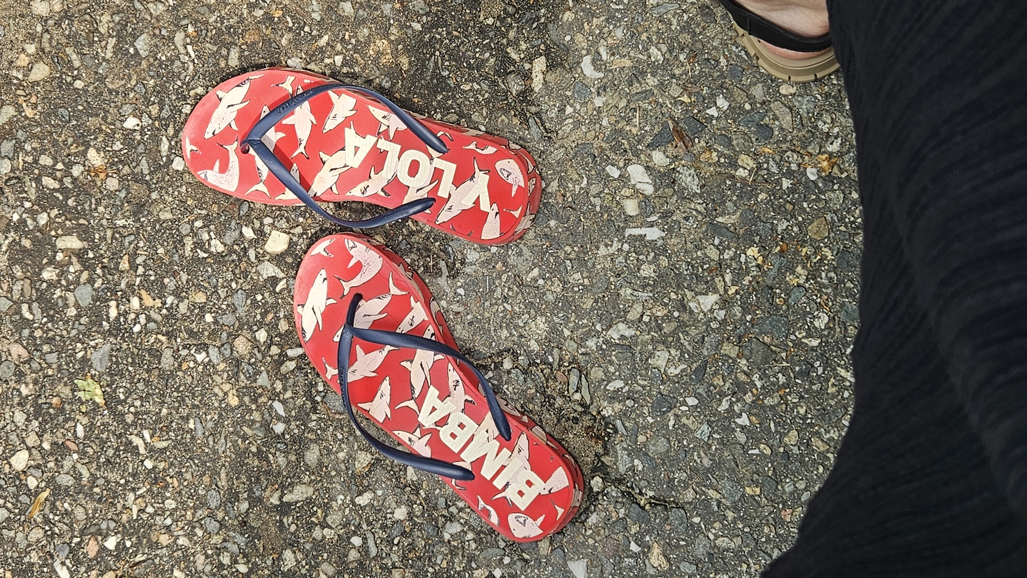 A pair of red flip-flops with a shark pattern and blue straps are lying on a gravelly asphalt surface. The left flip-flop has the word "BIMBA" printed in white letters on its sole. The right flip-flop has the word "LOLA" printed in white letters on its sole. A person's foot wearing a sandal is visible in the lower right corner of the image, and dark fabric is visible in the lower left corner. The location is the city of Dyurso, Russia.