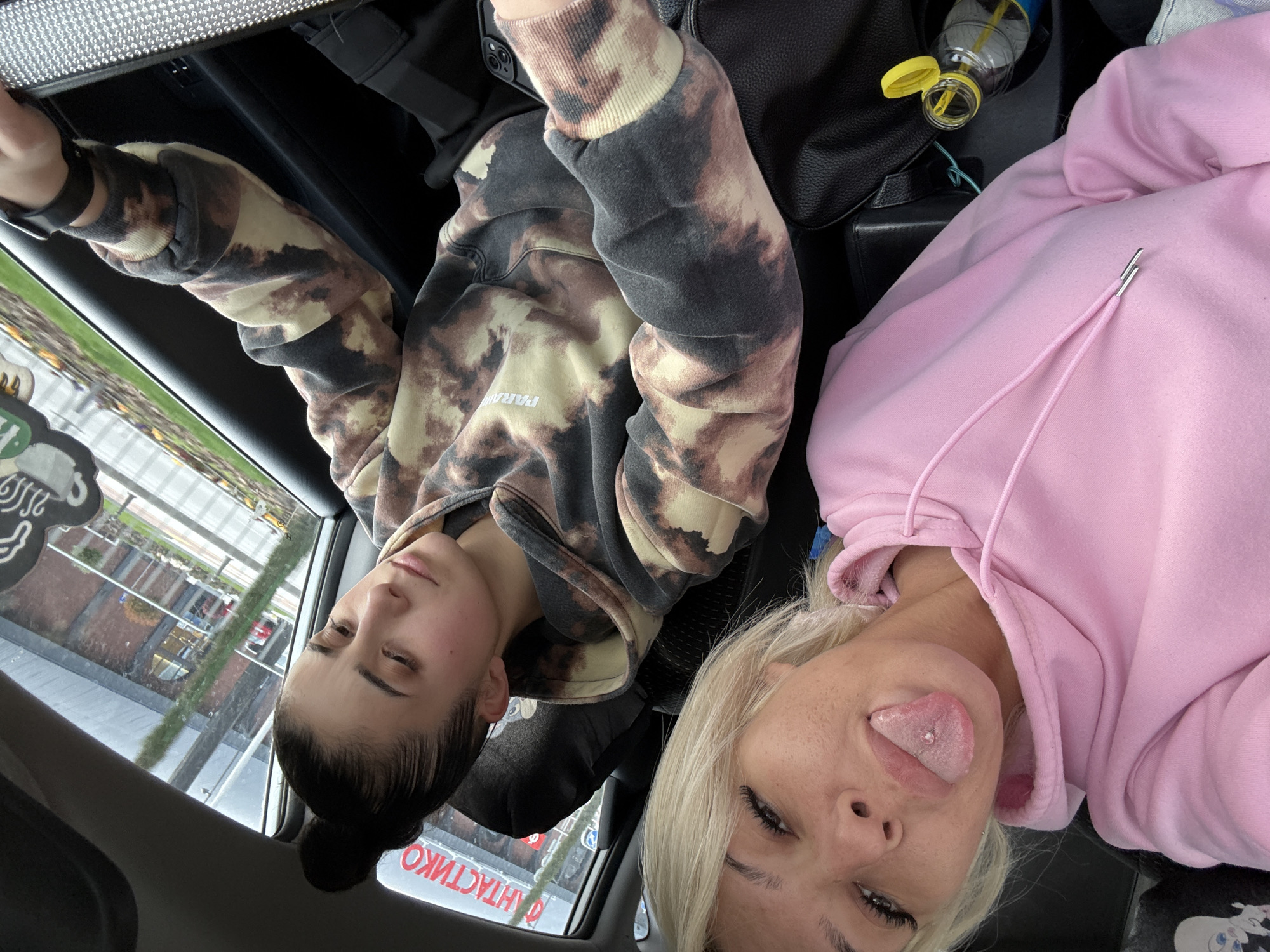 Two women are seated in a car. One woman, with dark hair pulled into a bun, is wearing a tie-dye hoodie. The other woman, who has blonde hair, is wearing a pink hoodie and sticking her tongue out. The car appears to be stopped near a building with Cyrillic writing.