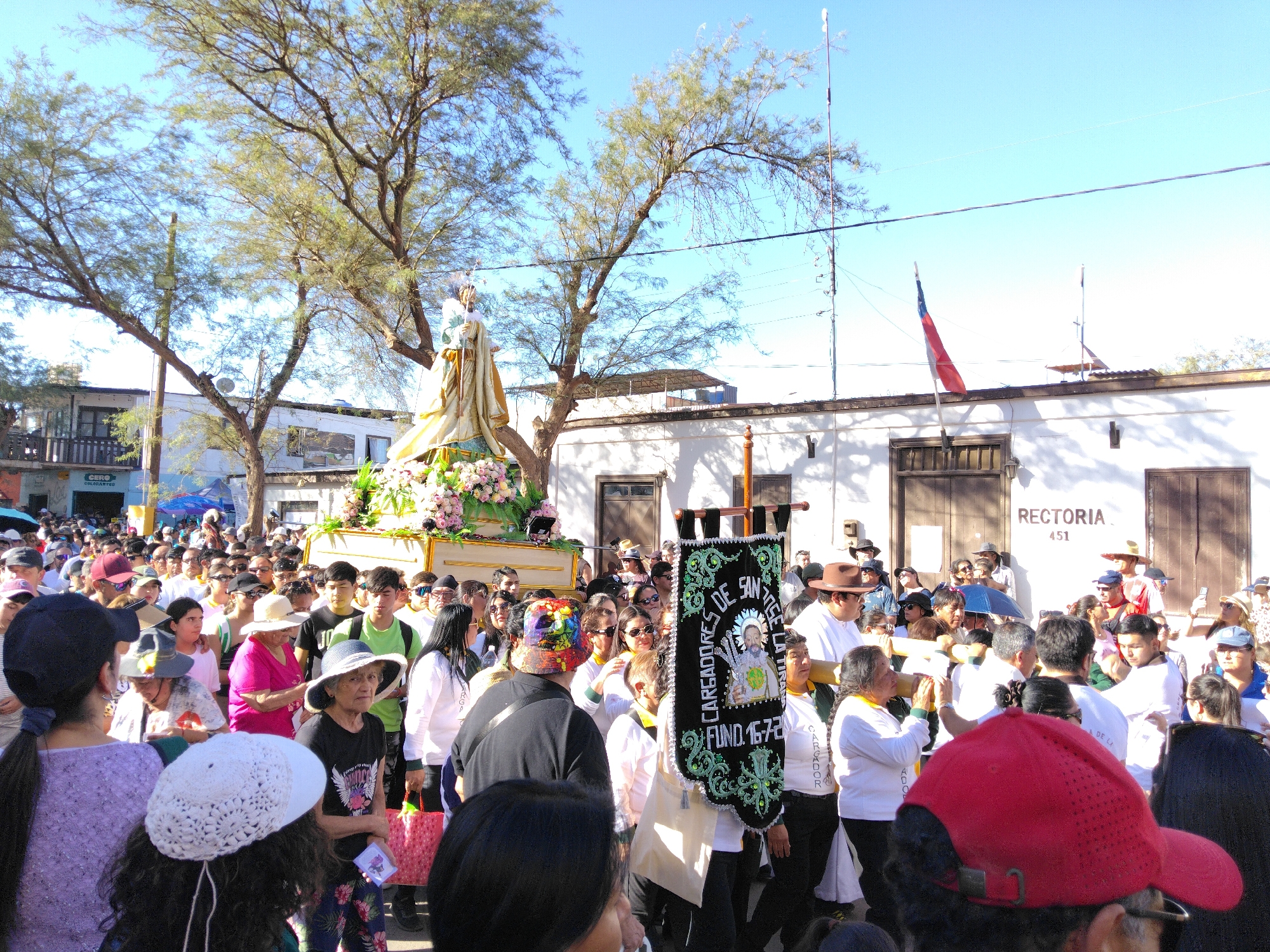 In Pozo Almonte, Chile, a religious procession is underway. A large statue of a saint, adorned with flowers, is being carried by a group of men. The statue is on a golden platform, which is being supported by large wooden poles. The men carrying the statue are wearing white shirts and black pants, and some are wearing hats. The procession is taking place on a street in the city, with buildings and trees visible in the background. The crowd of onlookers is diverse, with people of all ages and backgrounds. They are dressed in casual clothing, and many are holding cameras and cell phones to capture the event. The sky is clear and blue, indicating good weather. The overall atmosphere is festive and celebratory.