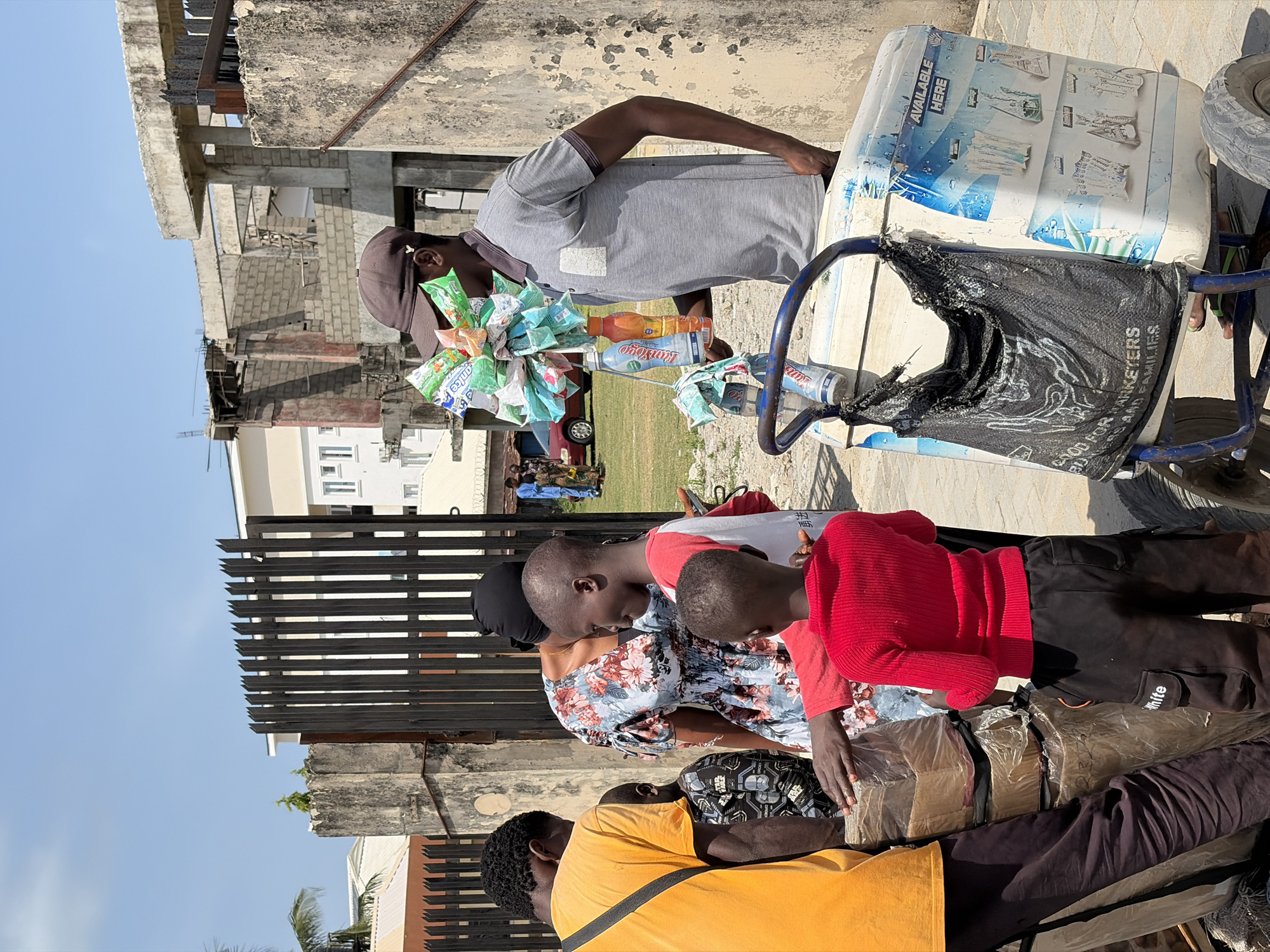 In Igboefon, Nigeria, a man wearing a gray t-shirt and a dark baseball cap stands behind a blue and white cart, examining a selection of packaged snacks and bottled drinks attached to the cart’s handle. The cart also has a black tattered bag hanging from it. The man is seemingly selling the contents of his cart to a group of three individuals. One person is wearing a yellow shirt, another is wearing a floral shirt and a black hijab, and a third individual is wearing a red sweater. The group is gathered around packages wrapped in plastic, which are stacked on a layer of clothing. A fence is between the people and a grassy area. In the background, a partially constructed building and a white building are visible. The sky is clear and blue.