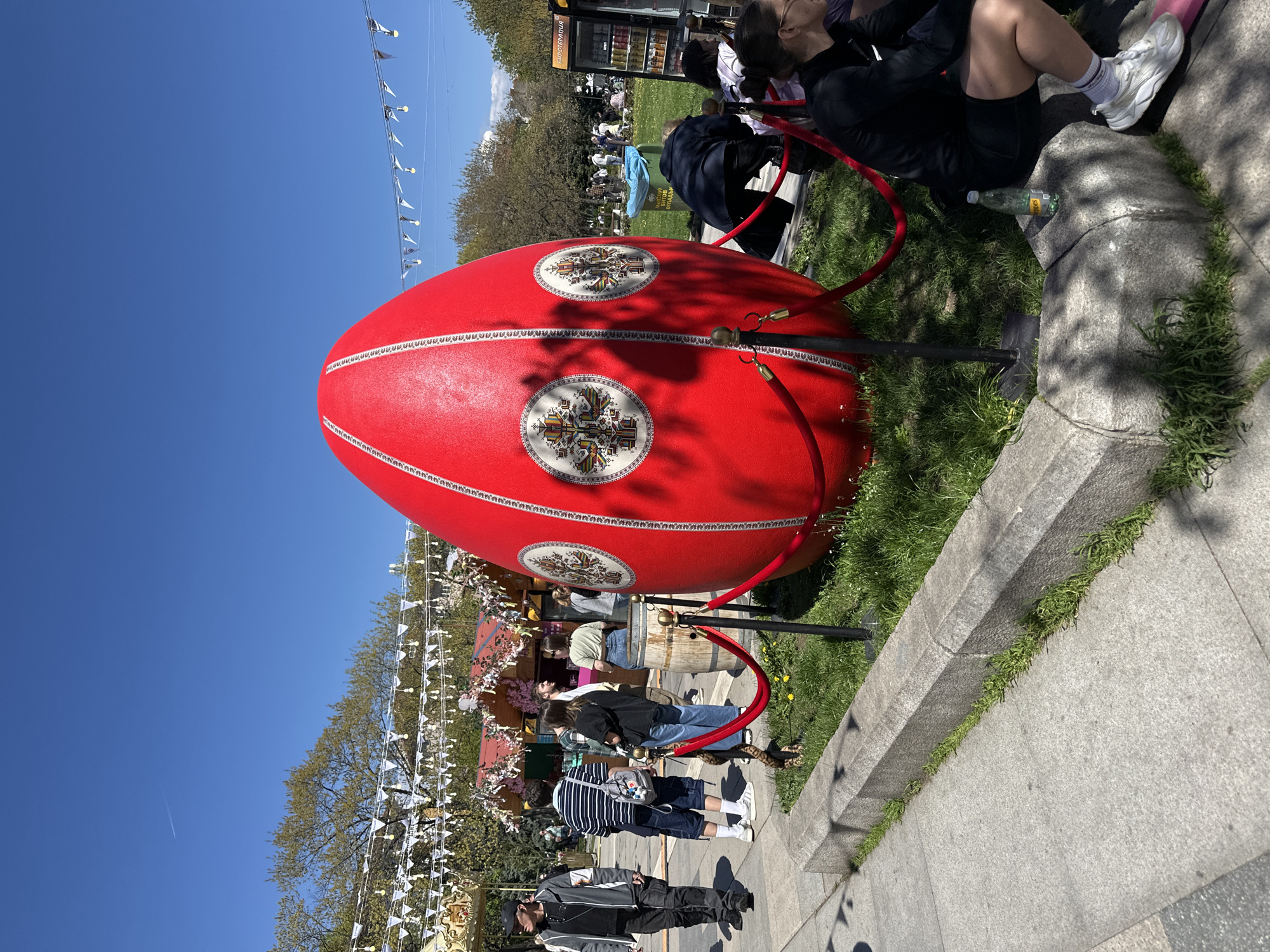 The image captures a large, red, decorative Easter egg, adorned with white and black emblems in circular frames, standing on a patch of green grass. The egg is partially surrounded by a red velvet rope barrier supported by black metal posts. Adjacent to the egg is a raised concrete sidewalk with small patches of grass growing along its edge.
Several people are visible in the background. Some are sitting on the grass near the egg, while others are walking along a path lined with decorated booths and white triangular pennant flags hanging overhead. A drinks vending machine is situated further back. The clear blue sky suggests it is daytime, and the bright sunlight indicates fair weather.
The scene is likely set in a public space, potentially during an Easter celebration. This image is of an Easter egg display in Sofia, Bulgaria.
