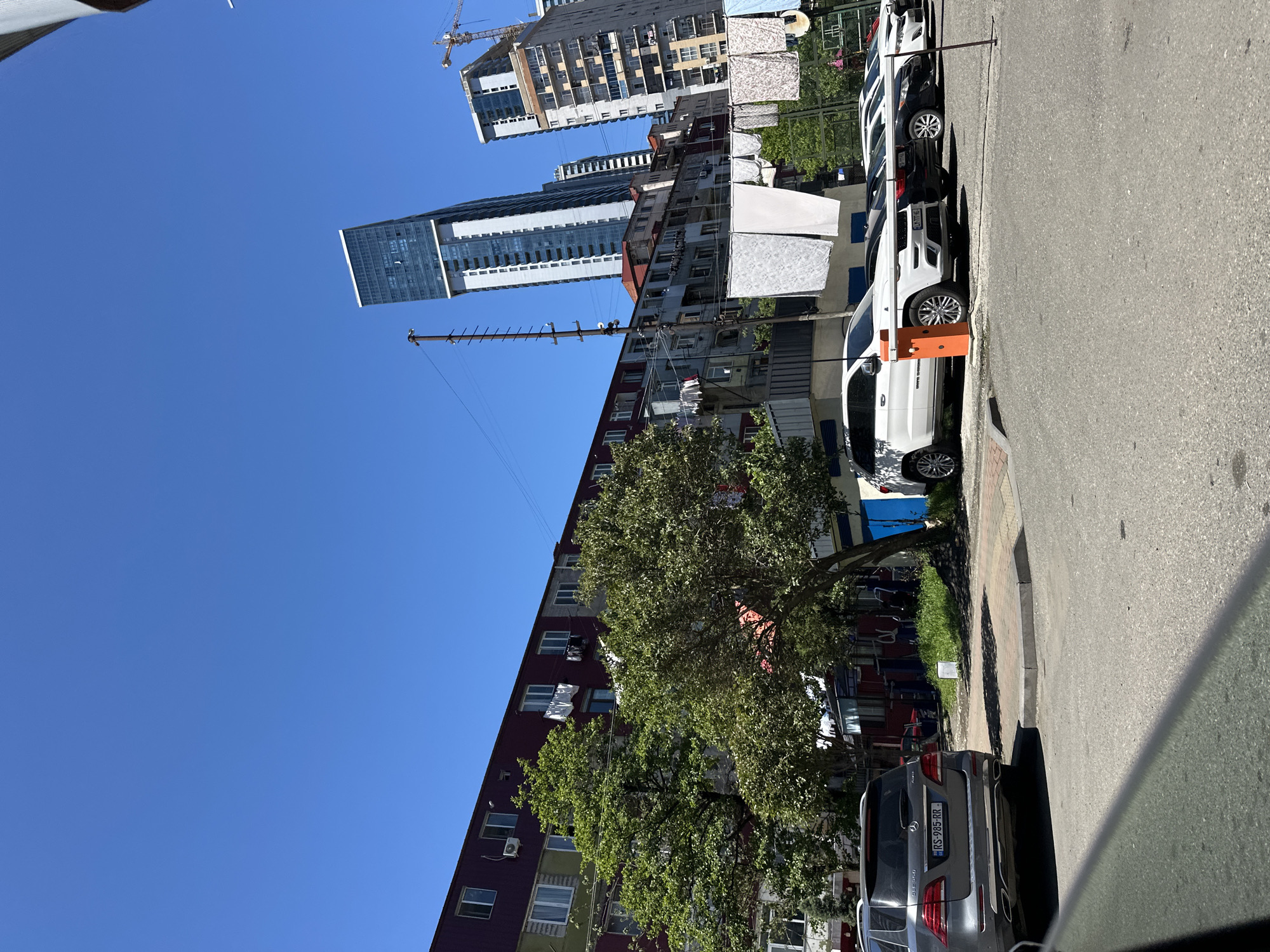 The image shows a street in Batumi, Georgia, with a view towards high-rise buildings. On the right side of the street, several cars are parked. The first car is silver with a license plate starting with "RS-985-RR". Next to the parked cars is a sidewalk and a row of buildings. The first building is a mid-rise apartment block with laundry hanging on clotheslines. Behind that apartment block are newer, taller skyscrapers, including one with a crane on top. The sky is clear and blue.
