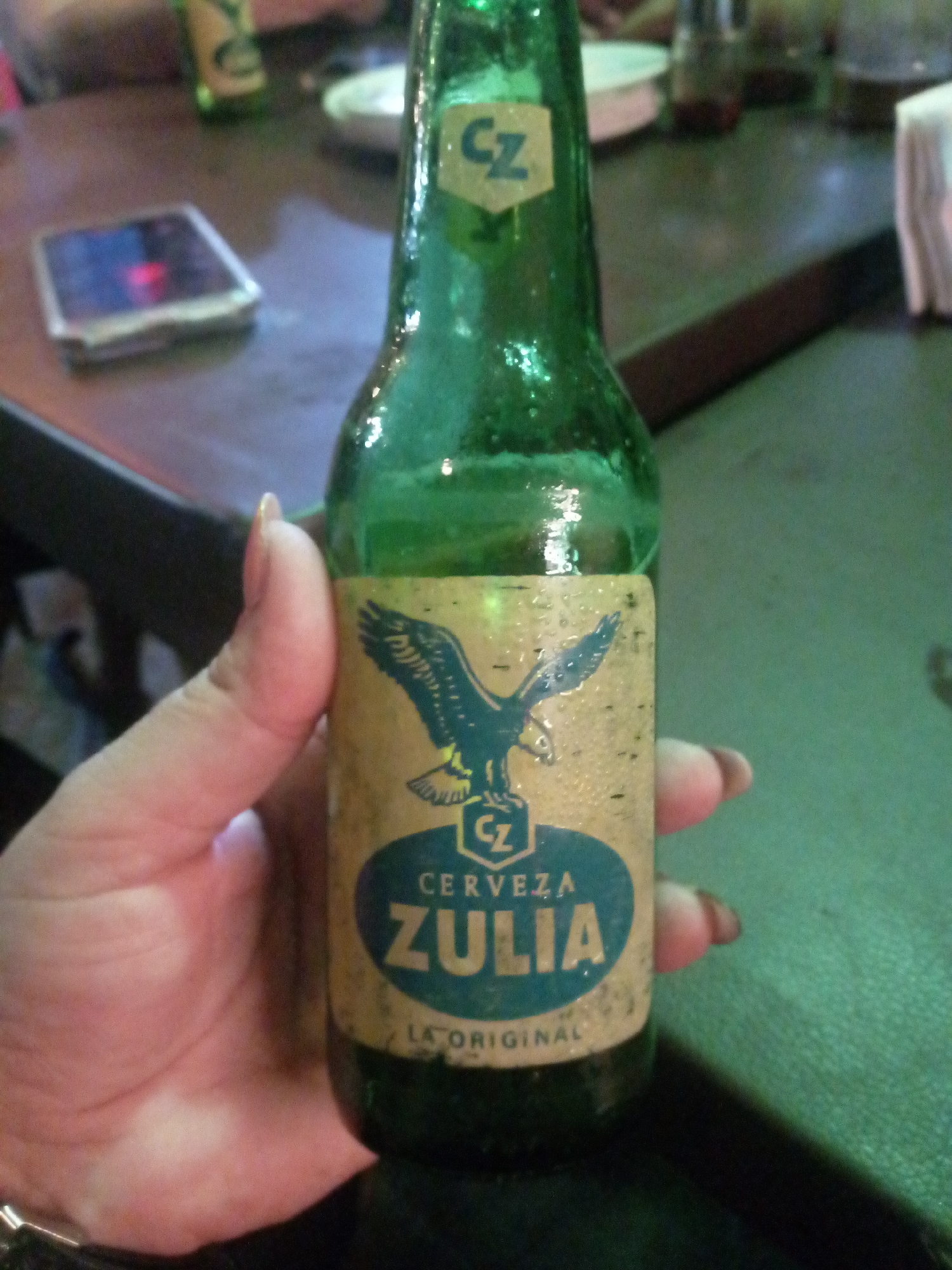 A person's hand holds a bottle of Cerveza Zulia beer. The green glass bottle is slightly wet with condensation. The label features an eagle above the beer's name in a blue oval. The words "La Original" are printed below the name. A CZ logo is visible on the neck and label. The background includes a table with a cell phone, another beer bottle, and napkins. The location is Juan Griego, Venezuela.