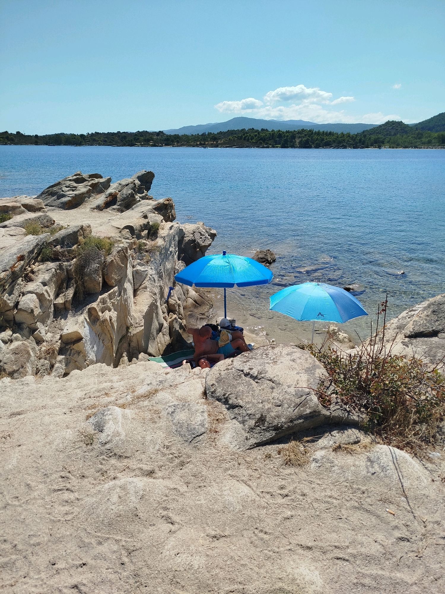 The image depicts a rocky coastline with clear blue water at Sithonia Municipal Unit, Greece. Two blue umbrellas are set up on the sandy shore, providing shade for two individuals who are lying down. The person on the left is partially obscured by the larger umbrella, while the person on the right is more visible, wearing a hat and resting. The surrounding environment is characterized by weathered rock formations, sparse vegetation, and the calm sea. In the background, a tree-lined shore stretches into the distance, with rolling hills and mountains under a clear blue sky with a few scattered clouds. The overall scene conveys a sense of peaceful relaxation on a sunny day.
