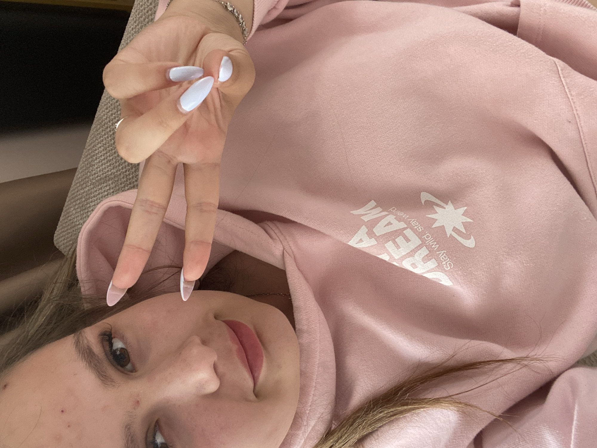 A person is seen in a selfie, lying down. The person is wearing a pink hoodie with white text. The fingers are raised in a peace sign. The person appears to be indoors, possibly on a couch. The city of Pletena, Bulgaria, is not evident in the image.