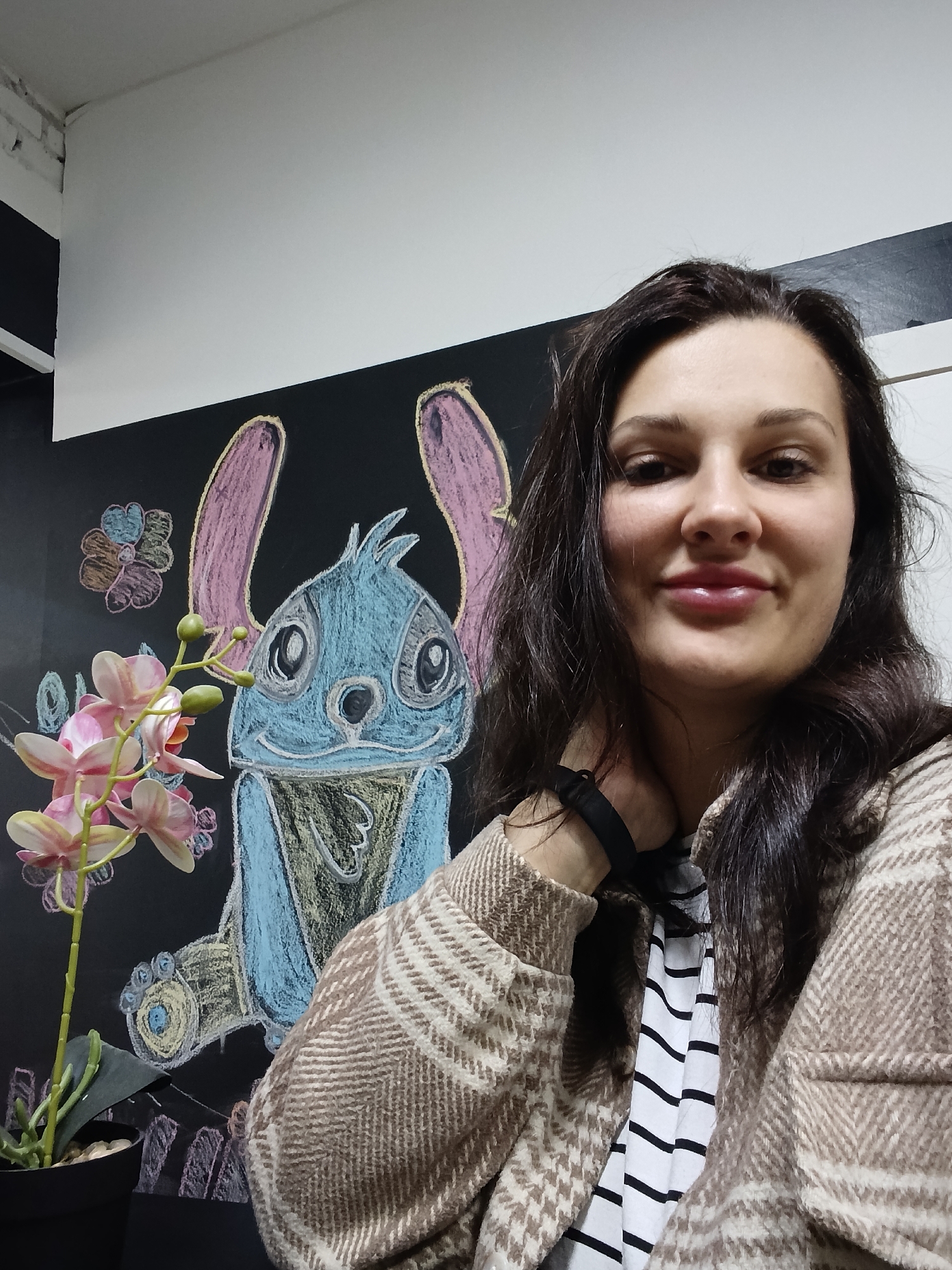 A woman with long, dark hair is captured in a close-up, chest-up shot, likely a selfie, in an indoor setting in Birobidzhan, Russia. She wears a warm, light brown and white plaid jacket over a black and white striped top. Her dark hair flows over her shoulders, and she has a gentle smile with slightly parted lips. Her left hand, with a black strap on the wrist, is resting near her neck/shoulder.
Behind her, a prominent feature is a black chalkboard wall adorned with colorful chalk drawings. The central drawing is a large, blue Stitch character with pink ears, detailed with blue, pink, yellow, and green chalk. Several smaller, multi-colored chalk flowers are drawn around Stitch. To the woman's left, a potted orchid plant with delicate pink and yellow flowers and green buds adds a touch of nature to the scene. Above the chalkboard, a plain white wall or ceiling segment is visible, with hints of brickwork in the top left corner, suggesting an interior space with a blend of textures. The lighting is consistent with an indoor environment. The woman appears relaxed and friendly, posing directly for the camera. There is no visible text in the image.