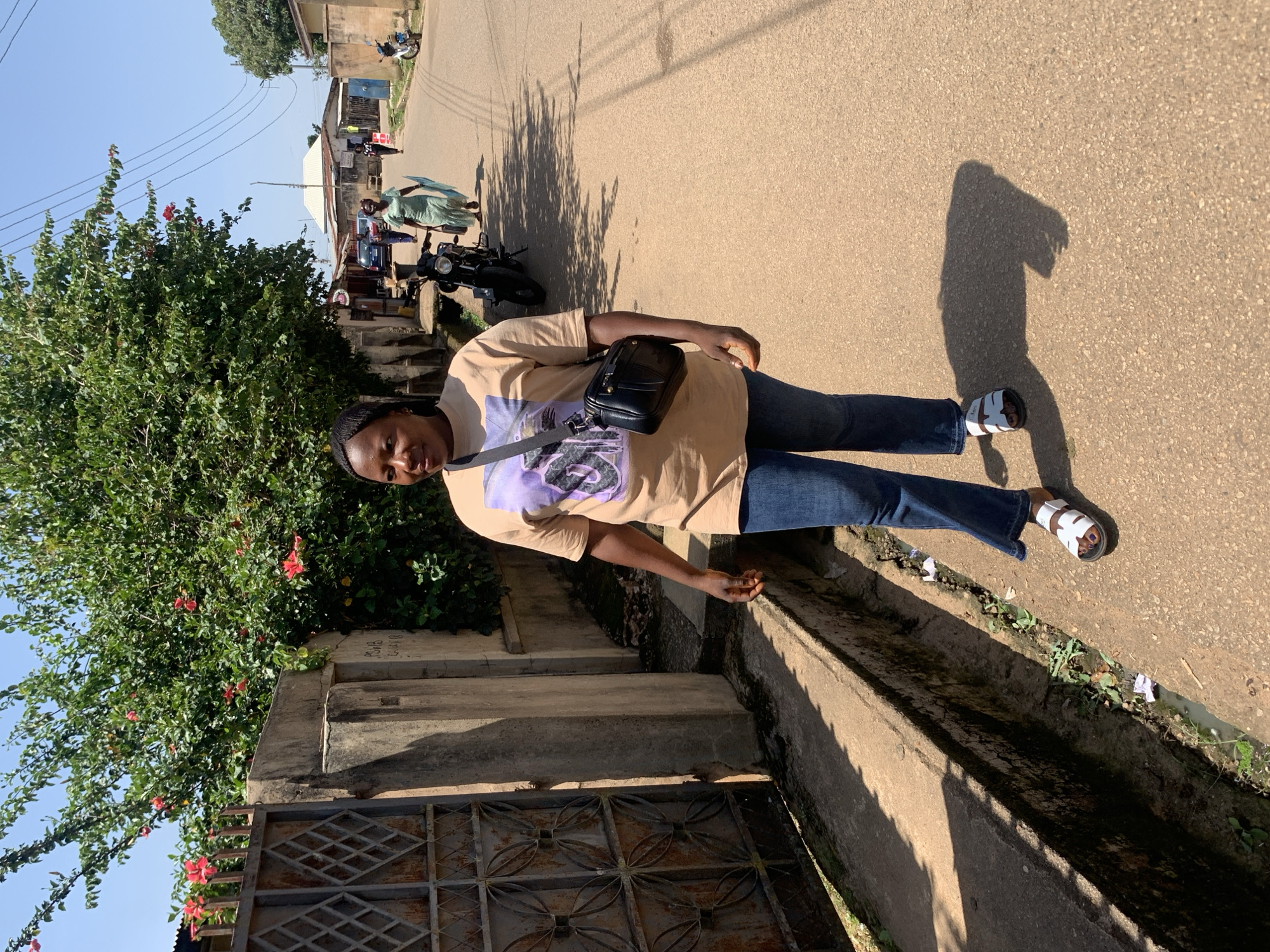 A woman walks down a paved street in Jos, Nigeria. She is wearing a light brown t-shirt with a graphic print, blue jeans, and white sandals. A black shoulder bag is slung across her chest. The street is lined with buildings and a large tree with red flowers. A metal gate is visible on the left side of the image. Other people and a motorcycle are in the background. The sky is blue and clear.