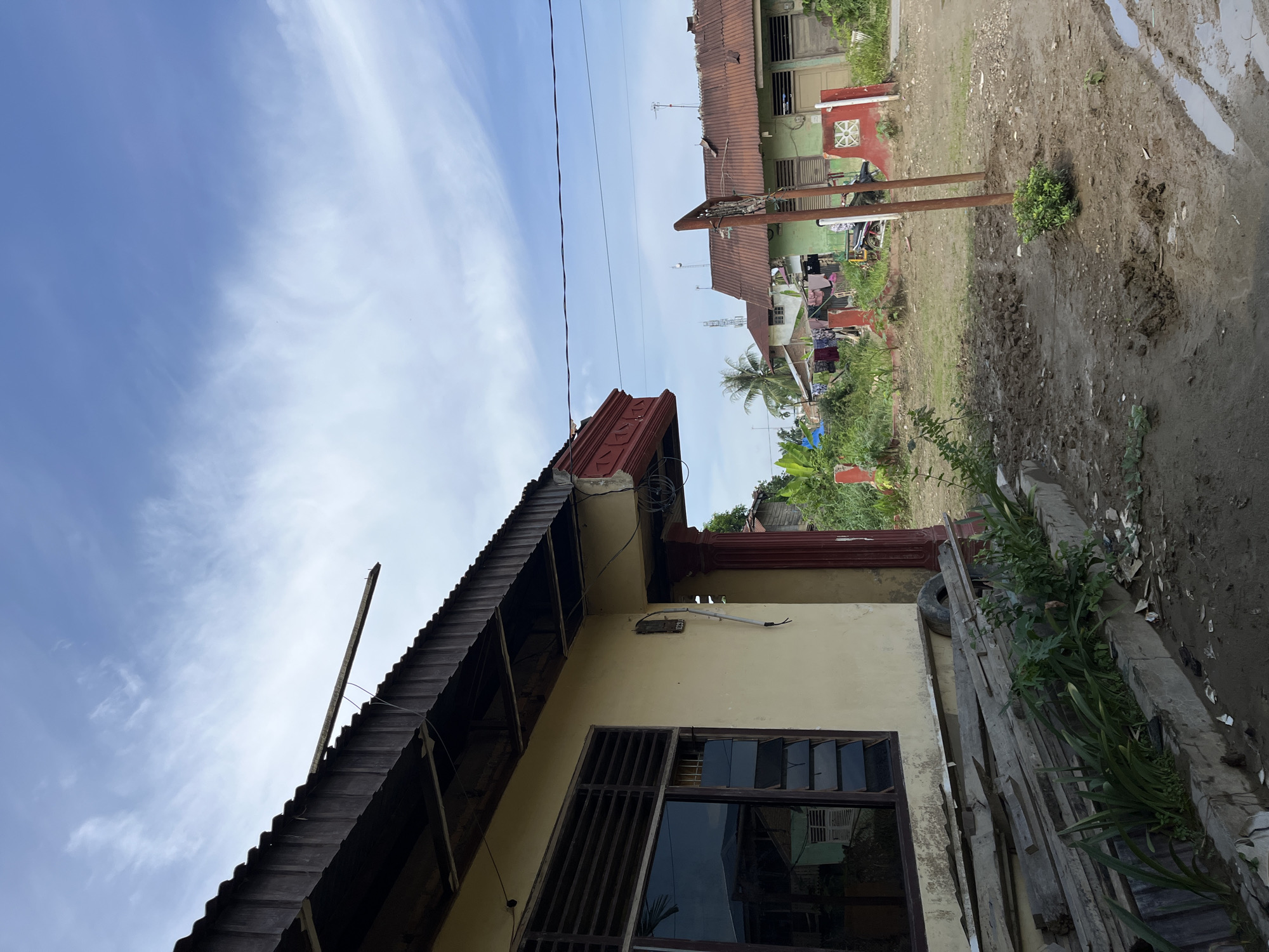 The image captures a street scene in Kota Kuala Simpang, Indonesia, under a partially cloudy sky. In the foreground, the side of a light yellow house is visible, featuring a dark-framed window with louvers and a corrugated metal roof. Piled wooden planks and some greenery are situated in front of the house. Beyond this, the scene opens up to reveal several other residential structures, some with red-tiled or rusted metal roofs, and lush vegetation. A basketball hoop structure stands in the middle ground, and a dirt path with muddy patches is present. The overall impression is that of a rural or semi-urban residential area.