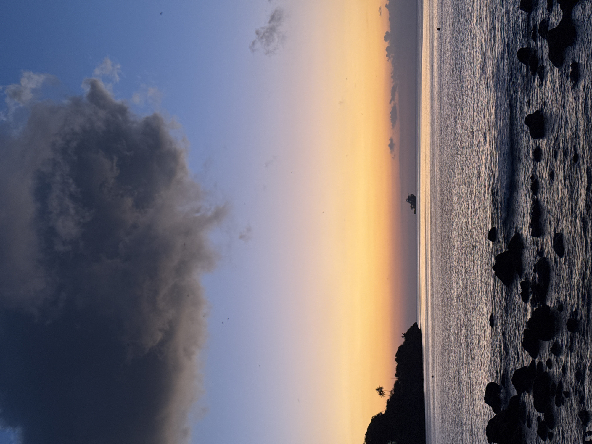 The image captures a serene coastal scene at dusk, presumably on Christmas Island, Australia, given the context provided. The sky is painted with a gradient of colors, transitioning from a deep blue at the top to soft purples and vibrant oranges near the horizon. A large, dark cloud dominates the upper left portion of the sky, contrasting with the lighter hues below.
In the distance, a ship is visible on the calm sea. The silhouette of a tree-covered headland, casting a dark shadow, juts out into the water on the left side of the frame. The foreground is occupied by dark, jagged rocks scattered along the shoreline, with gentle waves lapping against them. The overall mood is tranquil and picturesque, typical of a sunset over the ocean. There are no people or specific actions depicted in the image.