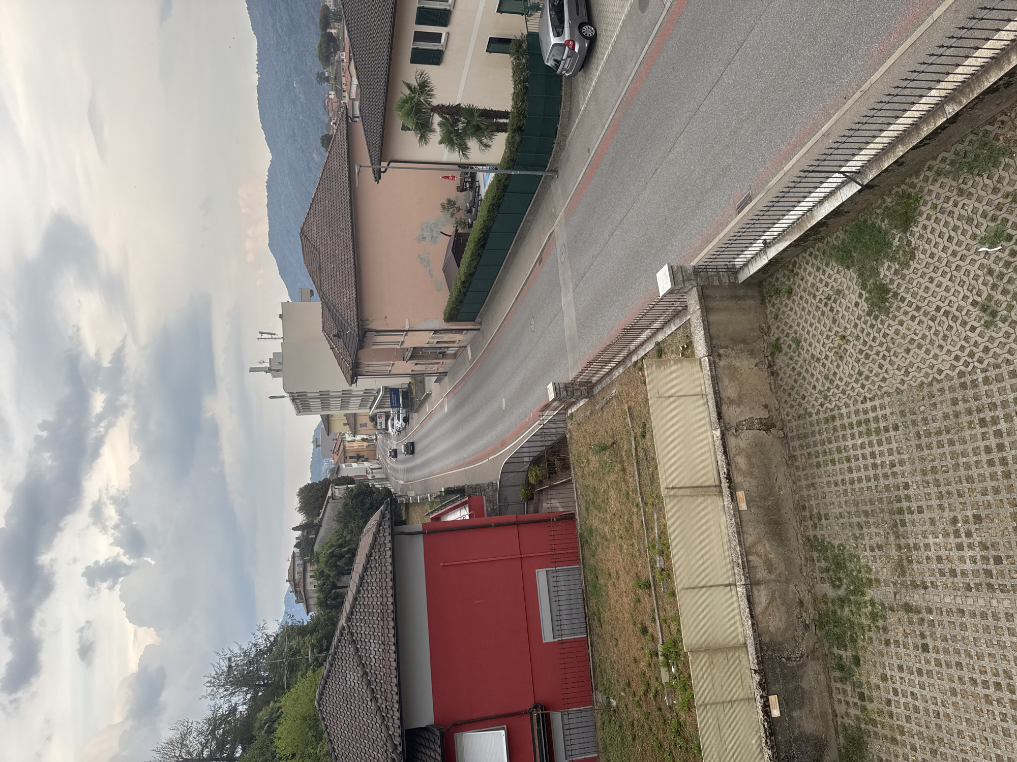 This image captures a view of a residential street in Balerna, Switzerland, under an overcast sky. In the foreground, a paved area with a grid pattern is visible, bordered by a concrete wall and an ornate metal fence. A section of dry, overgrown grass is situated between the paved area and the fence.
The street itself curves away from the viewer, lined on both sides by buildings and vegetation. On the left, a red and grey house with a tiled roof is prominent. The house features shuttered windows and a red facade. Further down the street, a light pink building with a tiled roof is visible, with a green hedge surrounding a small swimming pool area with loungers and an umbrella. Adjacent to this, a cream-colored building with green shutters stands.
Several cars are present on the street, including a silver hatchback parked on the right side. The street is marked with red lines. In the background, hills covered in trees rise up, indicating the surrounding natural landscape. Buildings with pitched roofs and some taller structures, including what appears to be an apartment building with visible antennas, are scattered along the street and on the hills. The overall impression is a quiet, residential area with a mix of architectural styles.