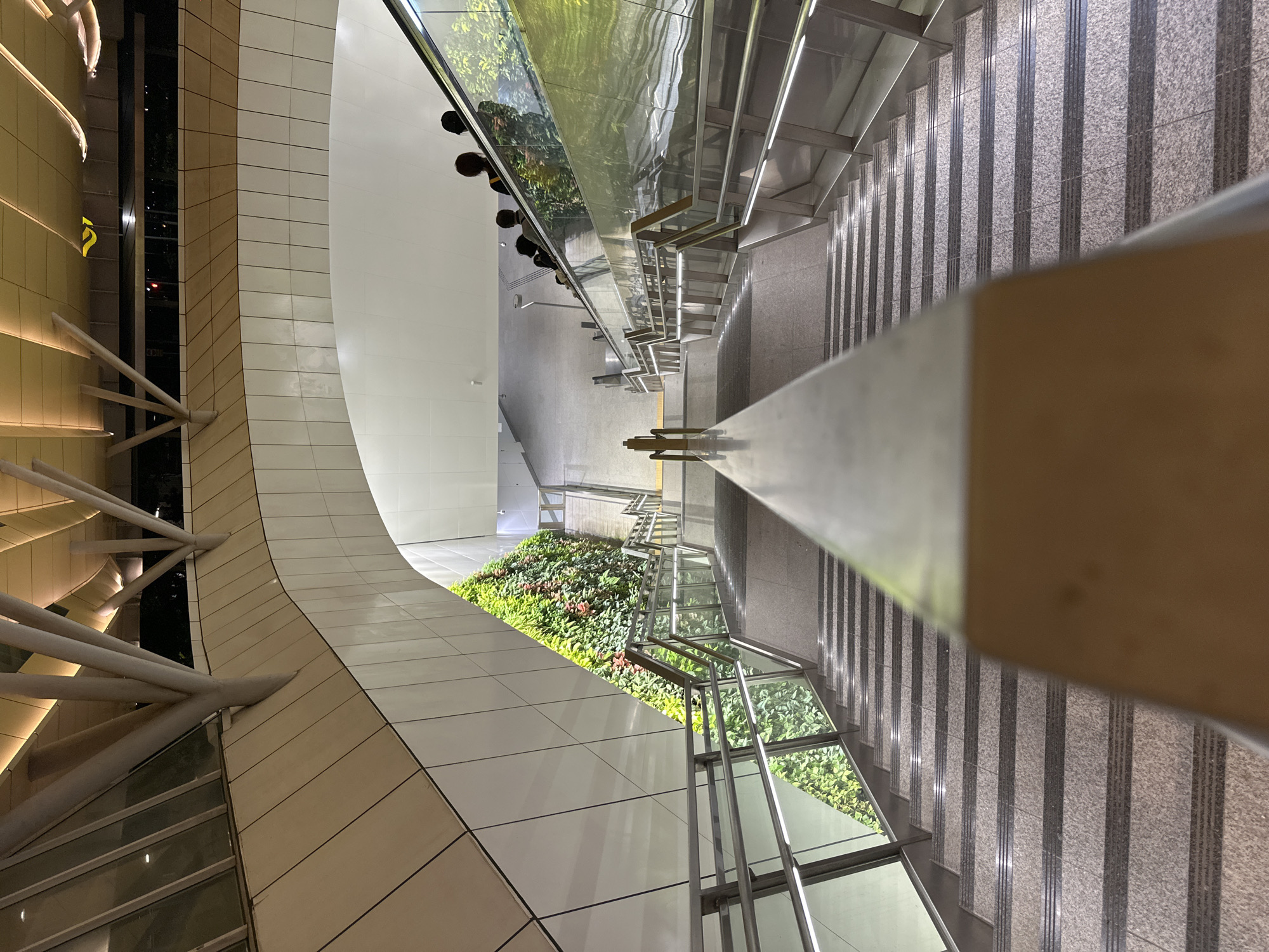 The image depicts a downward view of a modern escalator and adjacent stairs within an indoor urban setting, likely a transit station or shopping mall in Singapore. The escalator, with a glass railing, carries several individuals moving downwards. The stairs, made of speckled granite, are bordered by stainless steel handrails and glass panels. To the left of the stairs, a striking vertical garden wall is visible, featuring lush green foliage. The architecture above is characterized by curved white walls and a ceiling with integrated lighting, supported by cream-colored beams. The overall impression is one of contemporary design and urban infrastructure.