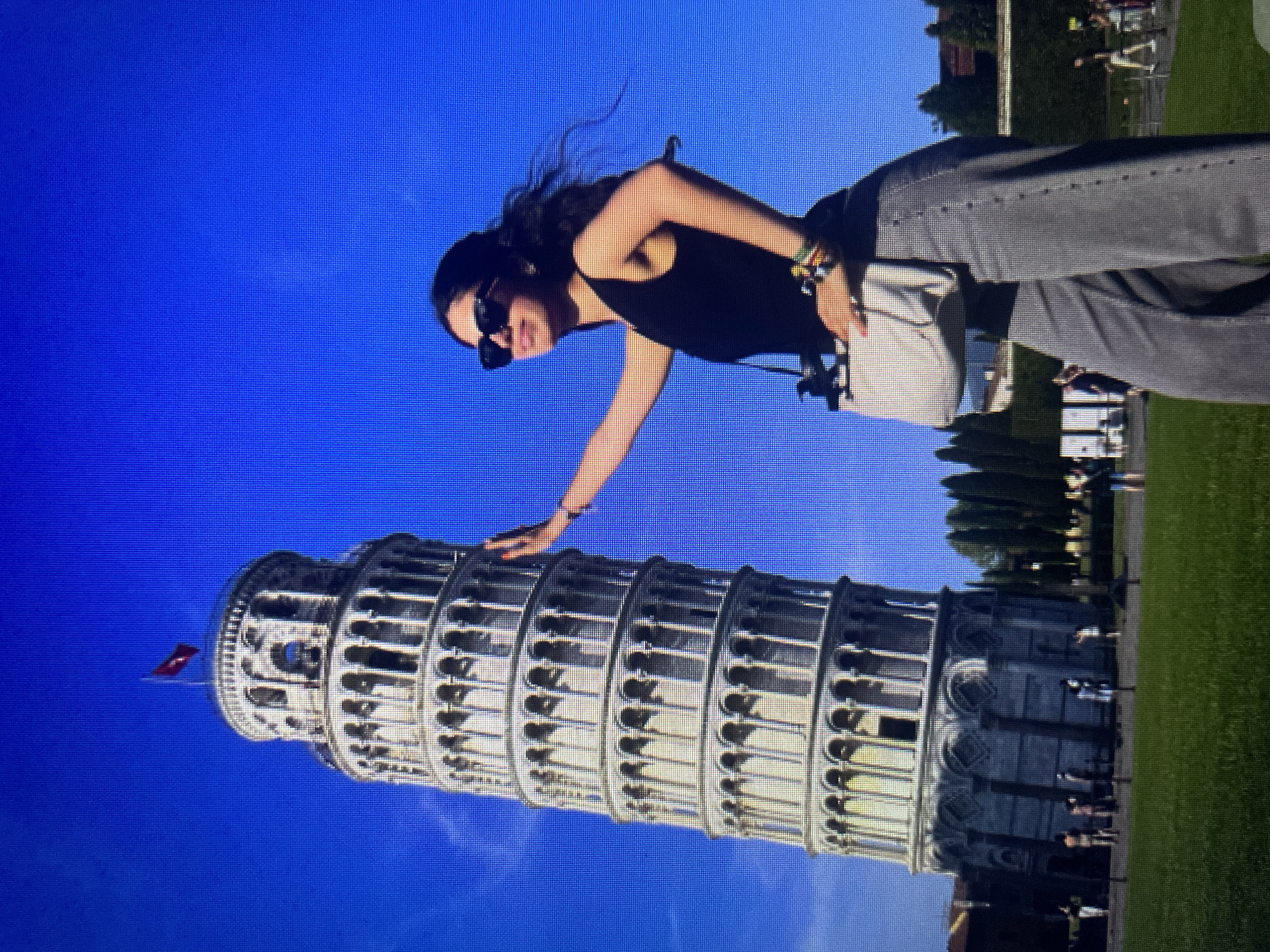 The image shows a young woman posing in front of the Leaning Tower of Pisa, Italy. She is wearing sunglasses, a black tank top, and gray wide-leg pants. She has a light-colored crossbody bag and several colorful bracelets on her left wrist. Her right hand is extended, appearing to touch the tower. The tower is a well-known landmark, and the sky behind it is a clear, vibrant blue. In the background, other tourists can be seen on the grass in front of the tower, and there are trees and buildings further in the distance. The scene is set on a sunny day, with the woman and tower casting shadows on the grassy area. The location is not in Kołobrzeg, Poland, but rather in Pisa, Italy.