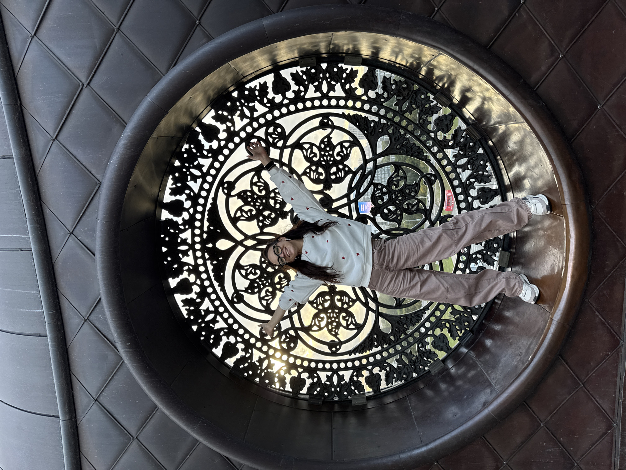 A young woman stands inside a large, ornate circular window with intricate metalwork, her arms outstretched. She is wearing glasses, a white sweater adorned with red hearts, and loose-fitting brown pants. The window appears to be part of a dark, textured architectural feature with a diamond-patterned exterior. The sunlight filters through the metalwork, creating a backdrop of a city skyline and trees. This location is the National Library of the Republic of Tatarstan in Kazan, Russia, which is known for its distinctive modern architecture featuring a large, symbolic "book" shape and this prominent circular window.