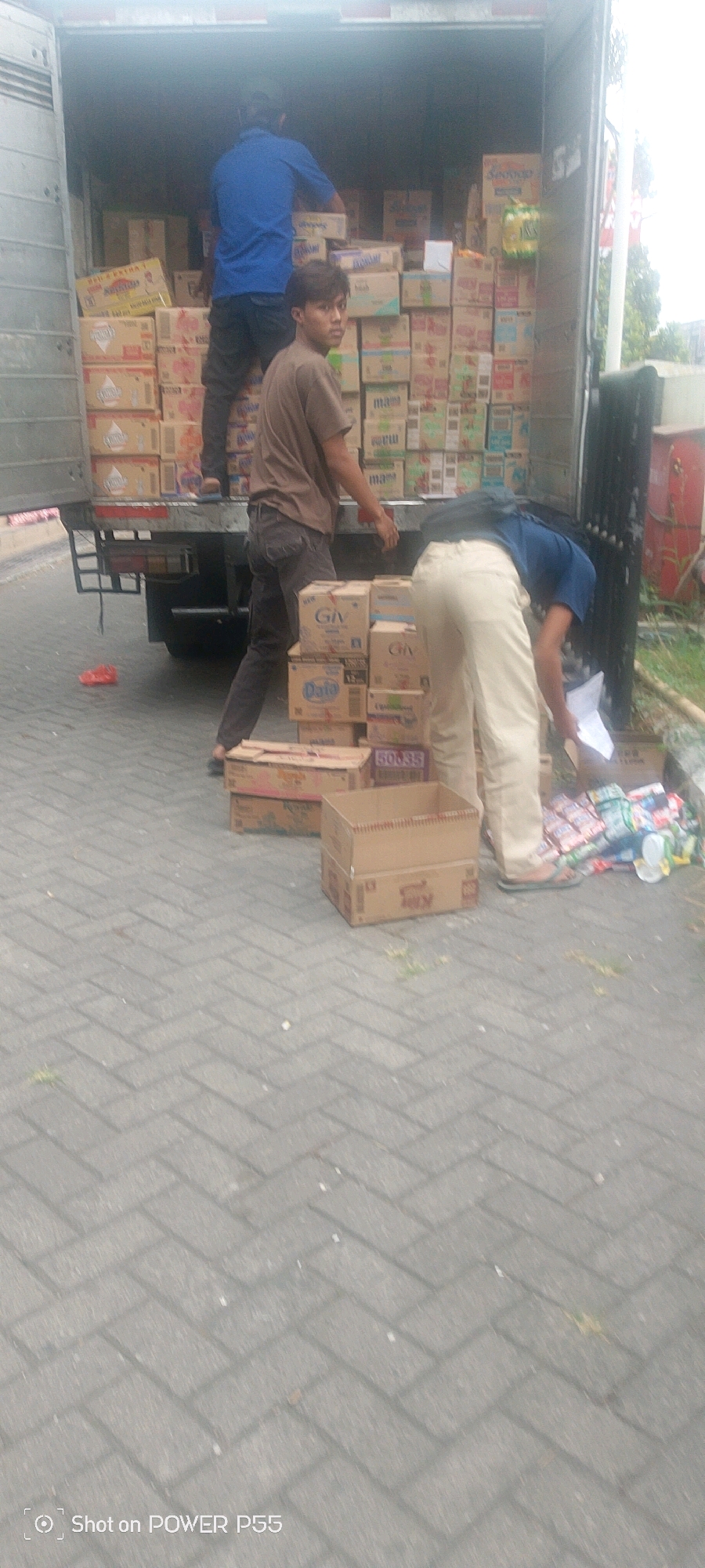 Two men are unloading boxes from the back of a truck. The truck is filled with stacks of boxes. One man, wearing a blue shirt and black pants, stands inside the truck, facing away from the camera, and appears to be sorting or moving boxes. The second man, wearing a brown t-shirt and dark pants, is standing closer to the front of the truck, looking towards the camera with his head tilted slightly. He has his hands down by his sides. A third man, wearing a blue shirt and light-colored pants, is bent over and looking at papers near a pile of smaller items and boxes on the ground. The truck is parked on a paved surface made of interlocking bricks. In the background, part of a red structure and some greenery are visible. A red plastic bag is on the ground near the truck's rear tire.