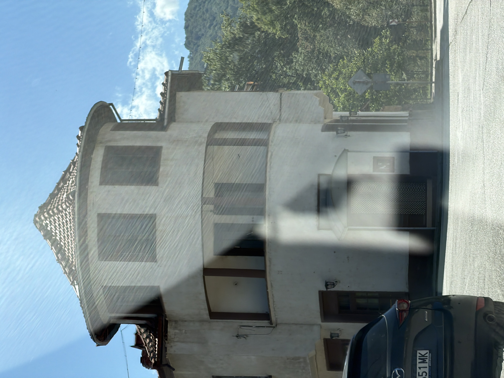 The image captures a slightly hazy view of a building with a curved facade and a tiled roof, situated in Beli Osam, Bulgaria. The building is a light color, possibly white or cream, with dark trim around the windows and doorways. It appears to have two stories. The windows on the upper level are square, while the ones on the lower curved section are arched. There is a street in the foreground with a dark SUV parked on the left side, its license plate reading "551 MK". Behind the building, lush green trees and a hillside are visible under a bright blue sky with some wispy clouds. The overall impression is a scene of a town or village setting, possibly on a sunny day, with the haze suggesting it could be warm or that the photo was taken through a window with some condensation or glare.
