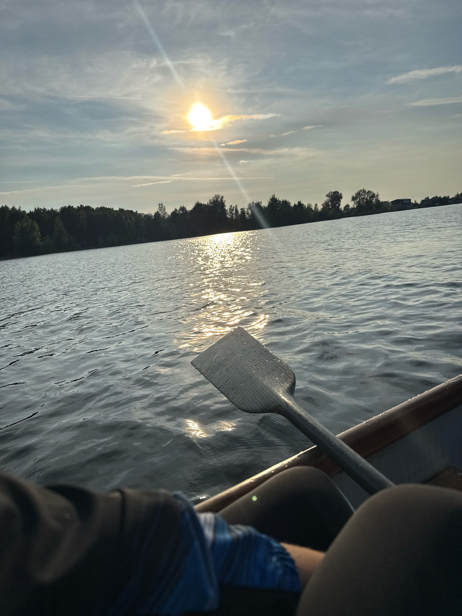 The image depicts a serene scene from the perspective of someone in a boat on a body of water, likely a lake or river. The sun is setting, casting a warm glow and reflecting brightly on the rippling water. A paddle lies in the foreground, partially submerged, indicating the boat is either stationary or moving gently. In the distance, a line of trees forms a dark silhouette against the sky, suggesting a natural landscape bordering the water. The sky is a mix of blue and grey clouds, with a distinct sunbeam cutting through the atmosphere. The overall impression is one of tranquility and natural beauty. This scene is very likely located on the Kama River within the city limits of Chaikovsky, Russia, given the geographical context of the city's location and its prominent riverfront.
