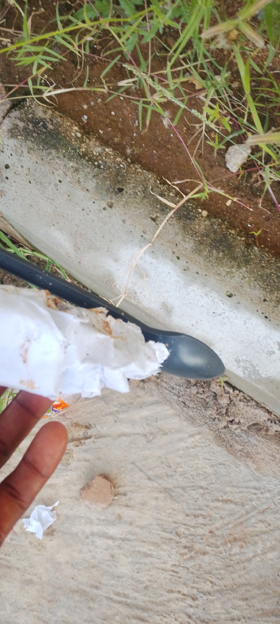 The image shows a close-up view of a hand holding a crumpled piece of white paper. The hand is dark-skinned, and the paper appears to be stained with dirt or mud. Behind the hand, a black object, possibly a spoon or a tool, is visible, partially submerged in dirt and resting on a concrete surface. The background consists of dry soil, green grass, and some small stones, suggesting an outdoor setting. A thin, dry twig or stalk is also present in the scene. The lighting suggests it could be daytime. The setting appears to be a natural or semi-natural environment, possibly a garden or roadside area in Jalingo, Nigeria. No text is visible in the image.