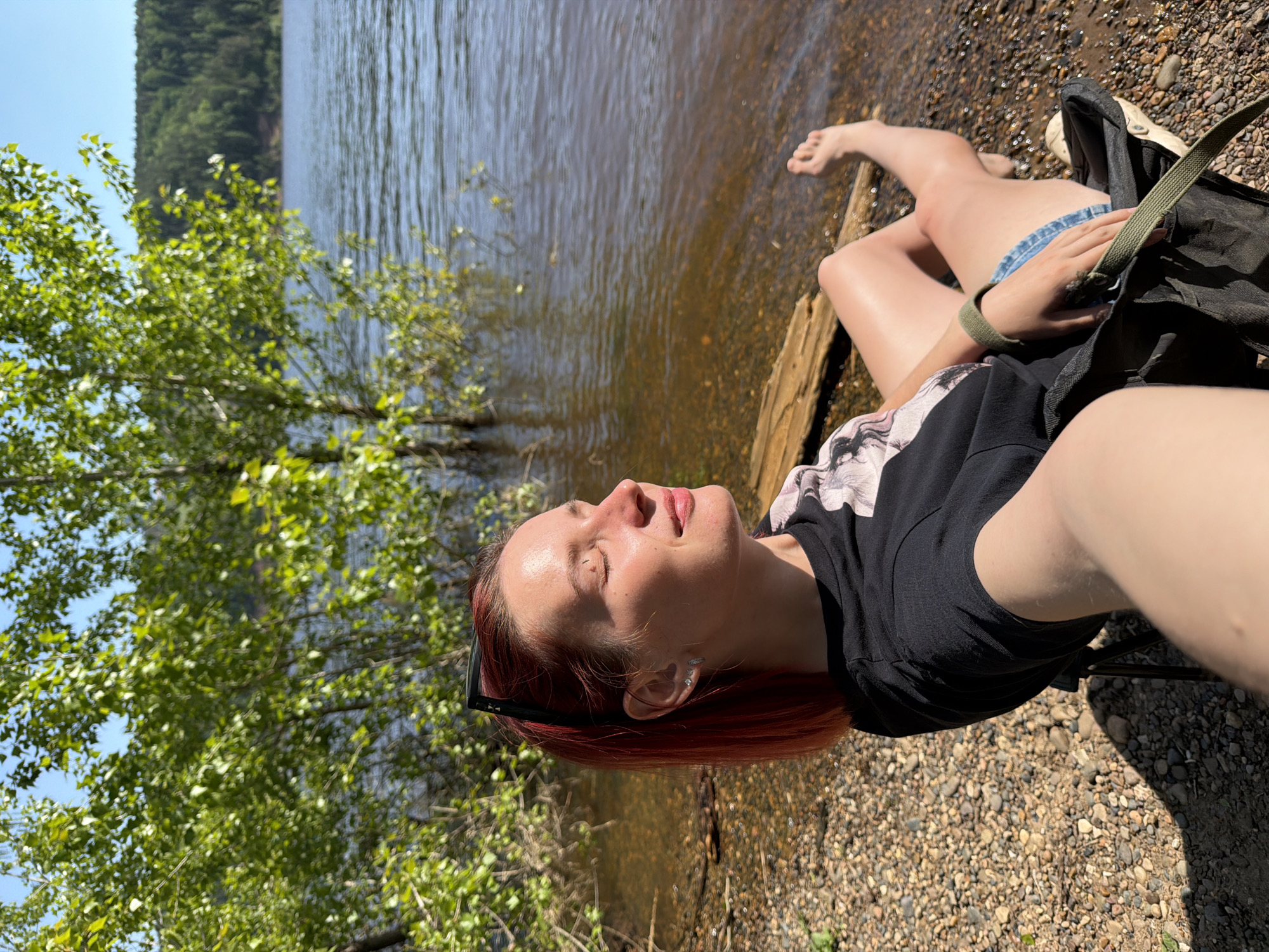 A woman is reclining on a rocky bank next to a body of water, possibly in Ольховка, Russia. She is lying on a log near the water's edge, with her feet pointing towards the lake. She wears denim shorts, a black t-shirt, and has reddish-brown hair. Her eyes are closed, and she appears to be sunbathing or relaxing. A black backpack rests near her legs. The lake or river stretches into the distance, with trees lining the opposite shore. The foreground is a pebbly beach with small rocks. The sun casts bright light on the scene.