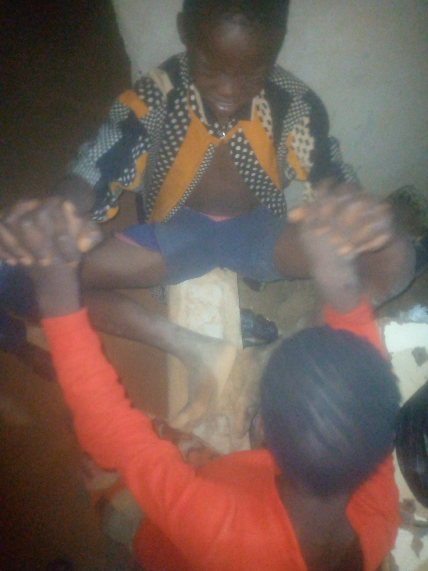 The image shows two individuals interacting in a dimly lit, indoor setting. The scene is somewhat blurry and grainy, suggesting low light conditions or a camera with limited resolution.
In the foreground, with their back partially to the viewer, is a person wearing a vibrant red long-sleeved shirt. Their head and upper back are visible, and their hands are raised and clasped together with the hands of the second person.
The second person is positioned in the background, facing towards the camera but looking downwards. They are wearing a distinctive patterned shirt featuring black, white polka dots, and yellow-orange sections. This person also wears dark shorts, and their bare legs are visible. A light-colored wooden or concrete block appears to be positioned between their legs, possibly used as a seat or an element of their activity. Their facial expression is unclear due to the blur and low light, but they appear focused on the clasped hands.
The primary activity involves both individuals with their hands tightly clasped together, suggesting a game, a shared ritual, or a close physical interaction. The setting appears to be a simple room, possibly with plain walls and an earthy or unpaved floor, indicative of a rustic or basic environment. No specific location or time of day can be determined definitively from the image, nor are there any visible texts. Emotions are not clearly discernible due to the image quality and obscured faces.