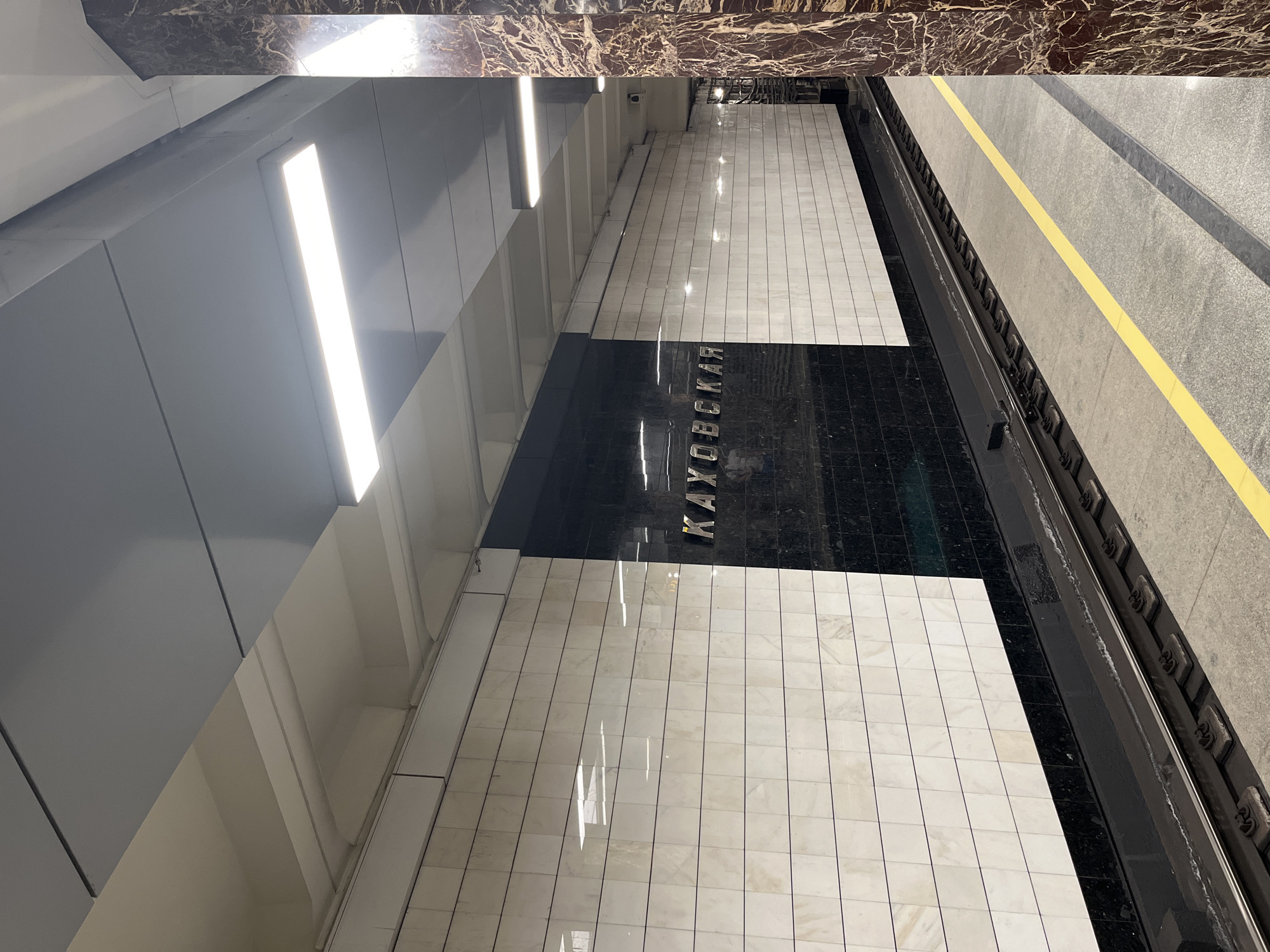 The image depicts the platform of the Kakhovskaya metro station in Moscow, Russia. The platform features a light-colored tiled floor with a dark-colored tiled area displaying the station's name in Cyrillic letters (КАХОВСКАЯ). The walls are a light gray color with linear lighting fixtures. A yellow safety line marks the edge of the platform, next to the train tracks. The area above the walls is decorated with dark, marbled tiles. The scene is devoid of people.