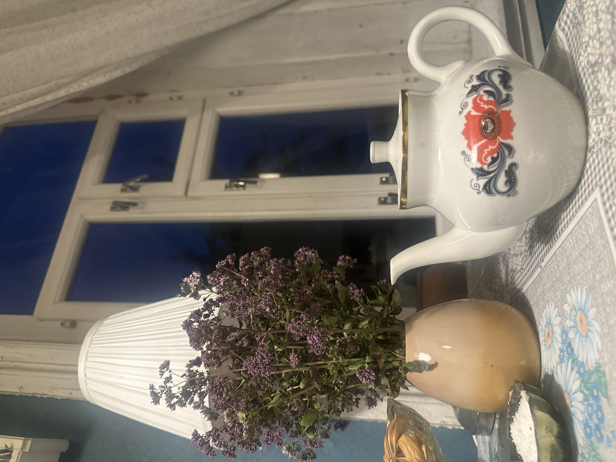 A still life scene is depicted indoors, likely in a home located in Staritsa, Russia. The background features a white-framed window with panes that reflect a dark blue sky, suggesting it is dusk or nighttime. A white, pleated lampshade is visible on the left, partially obscuring the window. In front of the lampshade, a bouquet of dried purple flowers with green stems is arranged in a light brown, ovular vase.
On a table covered with a patterned tablecloth, a white teapot with a gold trim and a decorative red and black floral pattern is prominently placed on the right side of the frame. To the left of the teapot, a small, dark bowl contains a white granular substance, possibly salt or sugar. Further to the left, partially visible, is a glass bowl filled with light brown, oval-shaped objects, likely cookies or crackers. The overall impression is a cozy, domestic setting.