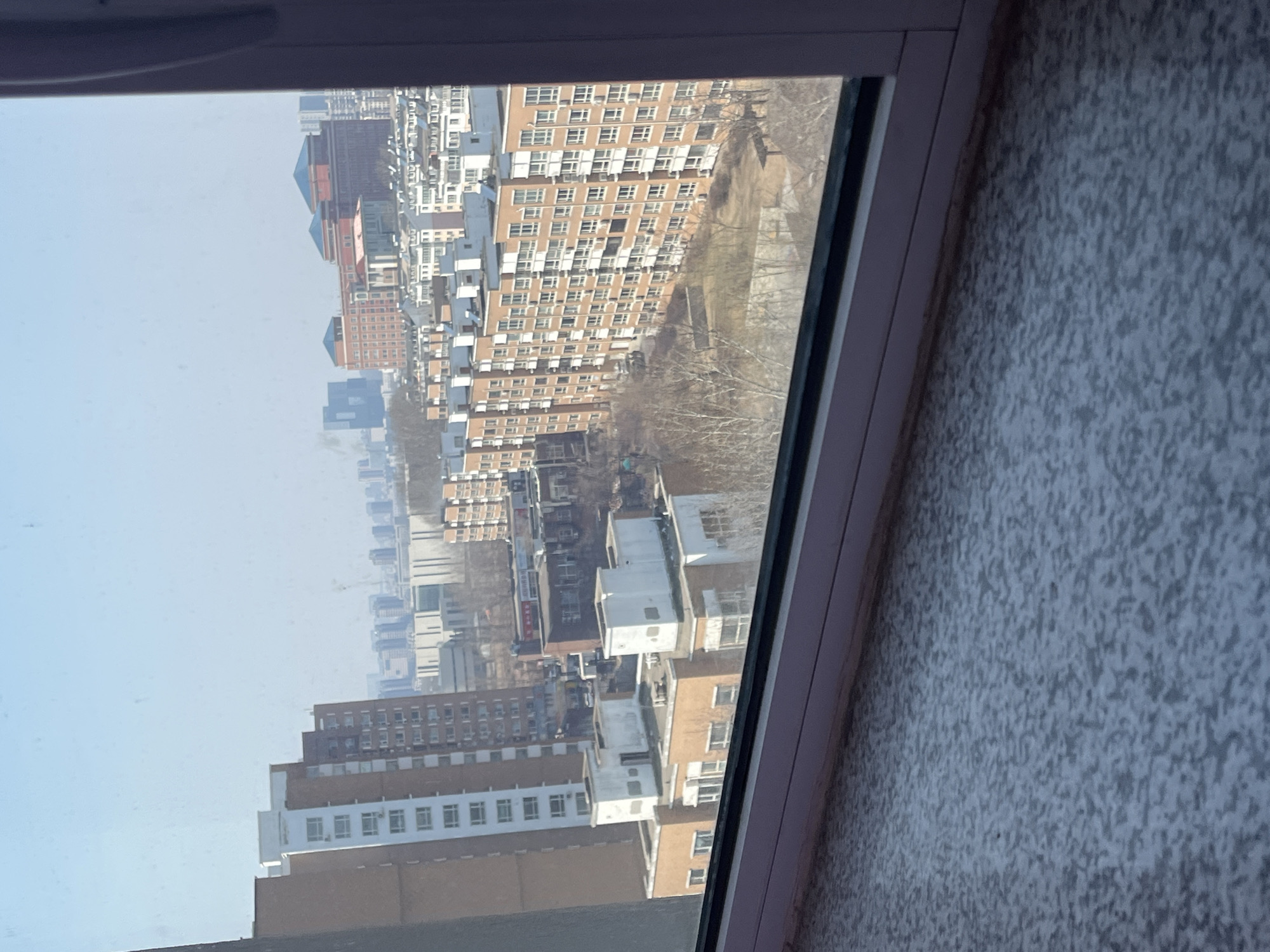 The image captures an elevated daytime view of an urban residential and commercial landscape in Changchun City, China, observed from a window. The bottom third of the frame shows a speckled light gray windowsill or table surface, bordered by a white window frame running horizontally across the lower edge and partially vertically on the right.
The sky above is a clear, pale blue. The midground is dominated by a dense arrangement of multi-story buildings. Several light brown/orange residential apartment blocks with white-framed windows and visible air conditioning units are prominent, particularly a long, uniform block extending rightward from the lower-middle. Many of these buildings feature flat rooftops with small, rectangular white structures, possibly utility enclosures. Interspersed among the buildings are patches of bare deciduous trees and sparse ground vegetation, indicative of a cold season.
In the central-background, a cluster of large, distinct brick-red buildings with dark blue, multi-pitched or pyramidal roofs is visible, suggesting institutional or significant commercial structures. Beyond these, the horizon reveals a distant skyline of numerous taller, modern high-rise buildings, slightly obscured by atmospheric haze. No specific individuals, vehicles, or dynamic actions are discernible.