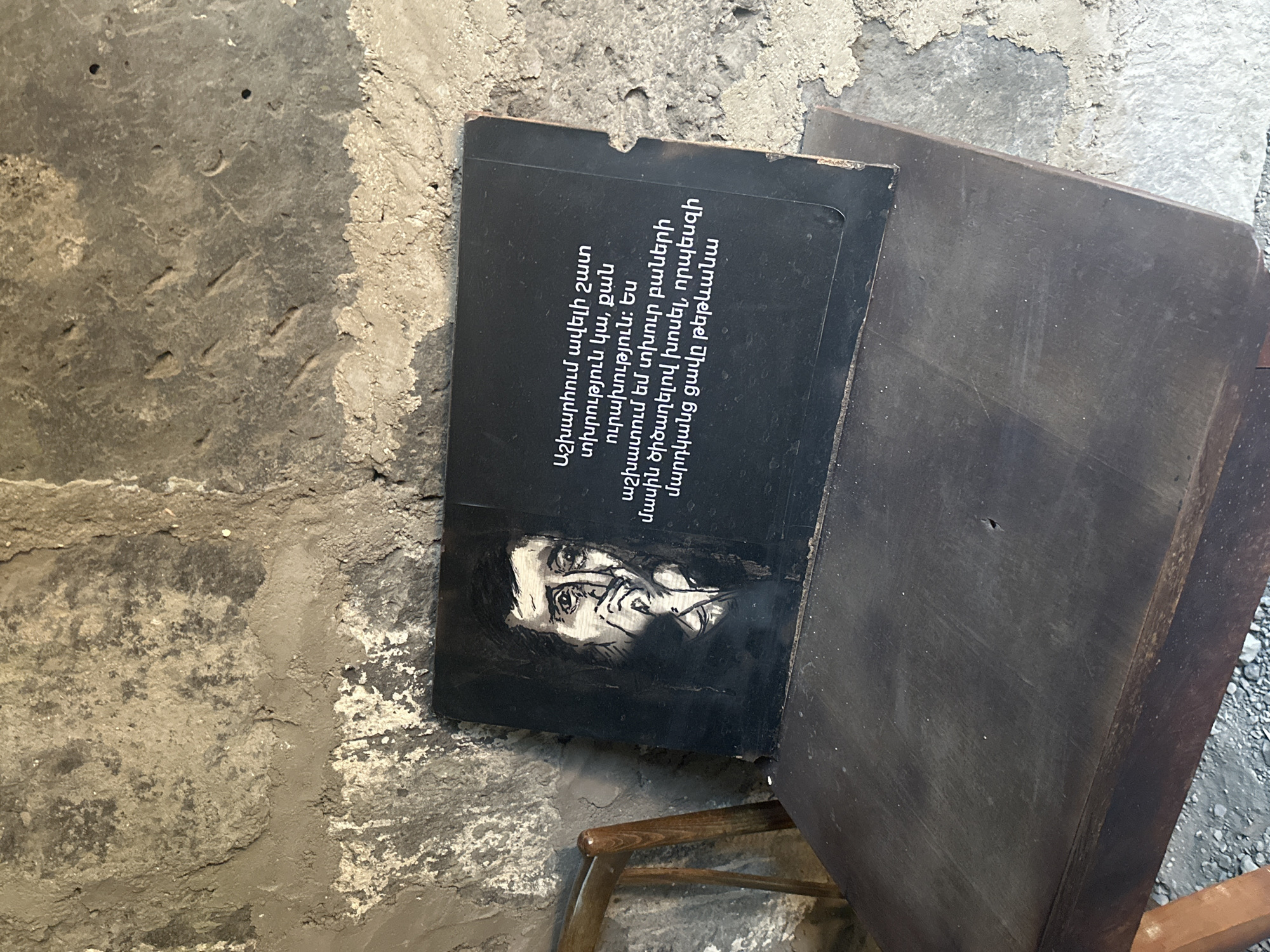 The image shows a black plaque with a sketch of a man's face on the left side and Armenian text on the right side. The plaque is placed on a dark wooden table, leaning against a rough, stone wall. The man's face is drawn in a dark, somewhat somber style, with his hand near his mouth. The text appears to be a quote or inscription, written in white. The environment suggests an indoor setting, possibly a historical site or a memorial, given the stone walls and the plaque's content. This scene is located in the "Old Gyumri" area of Gyumri, Armenia, specifically within the ruins of the St. John the Baptist Church. The church was heavily damaged in the 1988 earthquake and its remnants are preserved as a historical monument.